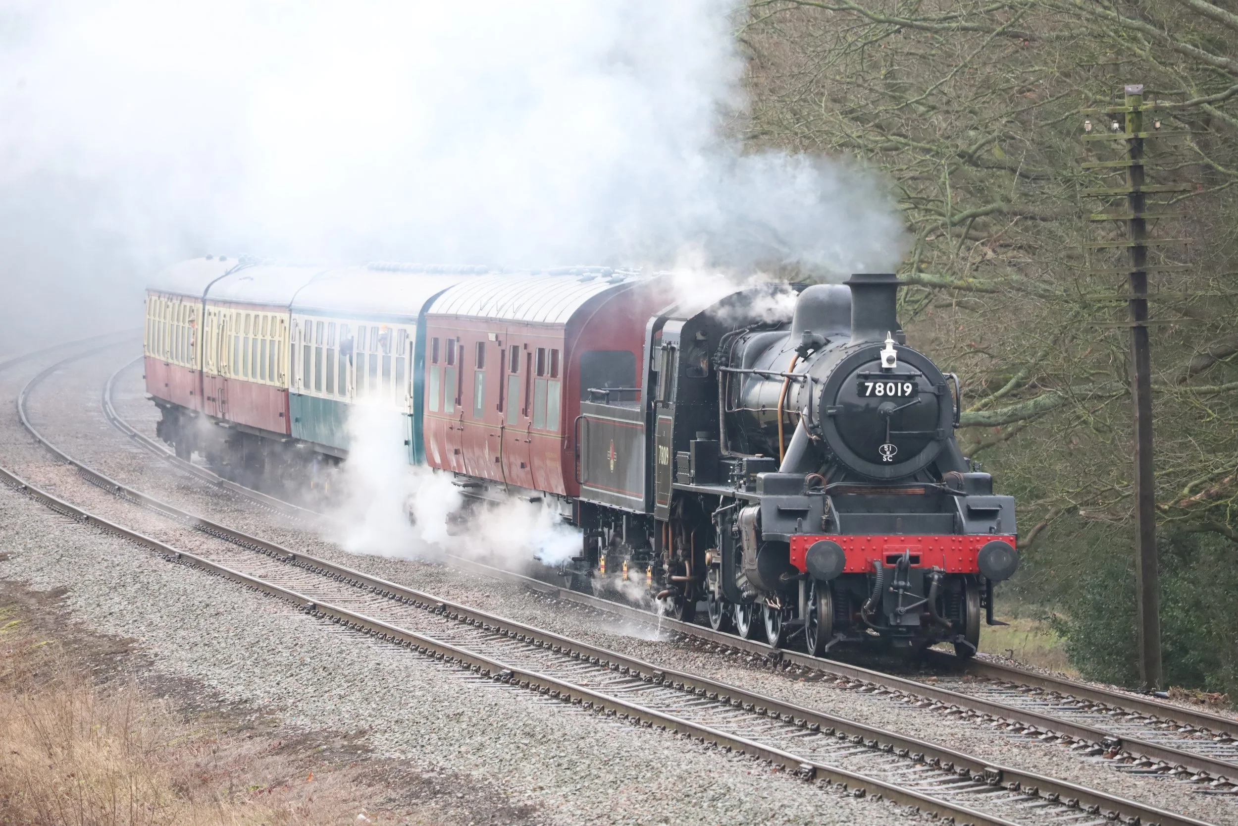A vintage black steam train with red accents and the number 78019 on the front, emitting white steam as it travels along railway tracks through a wooded area.