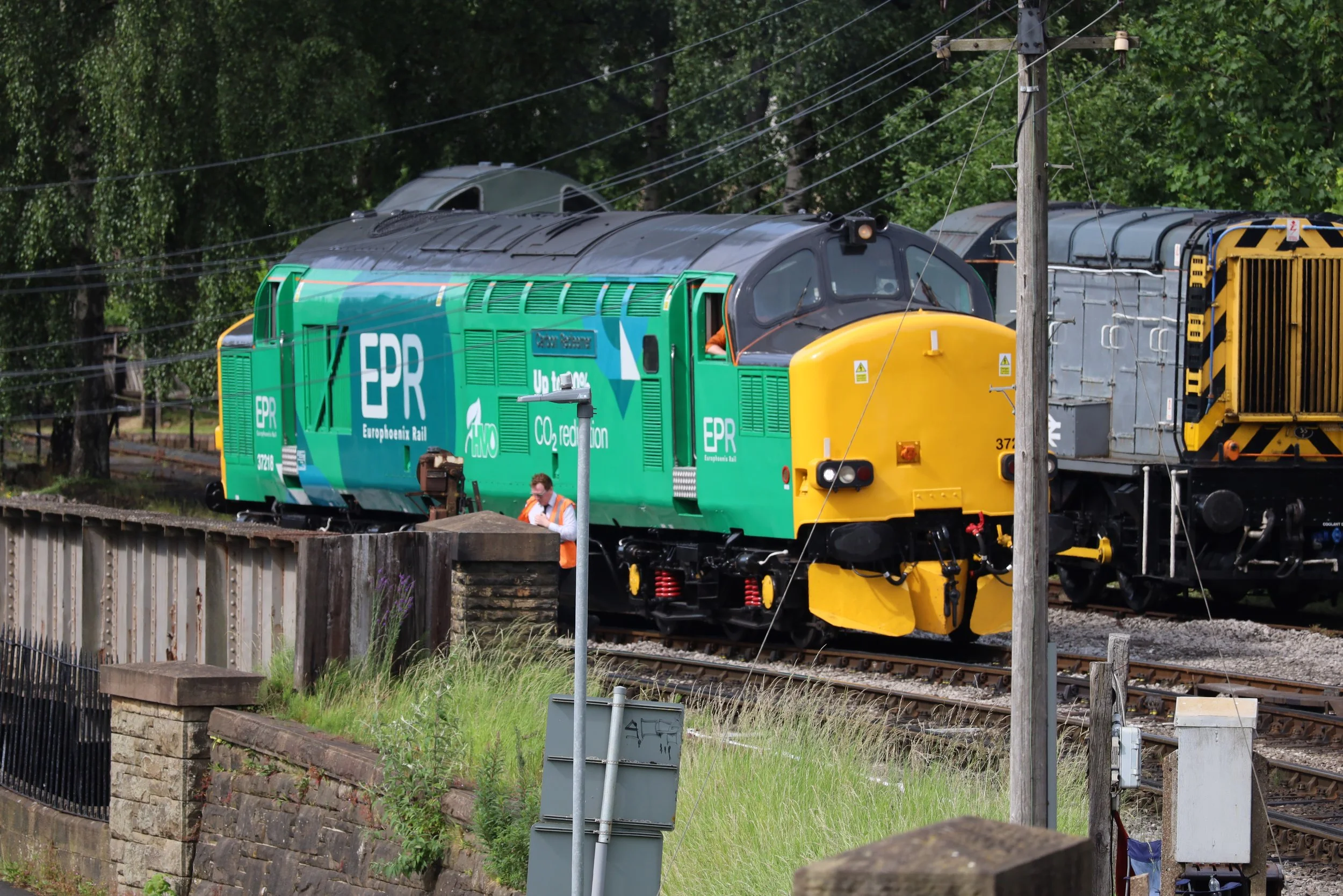 A green train engine on a railway track near a brick wall, with a man wearing an orange vest and face mask standing beside it, and trees and power lines in the background.