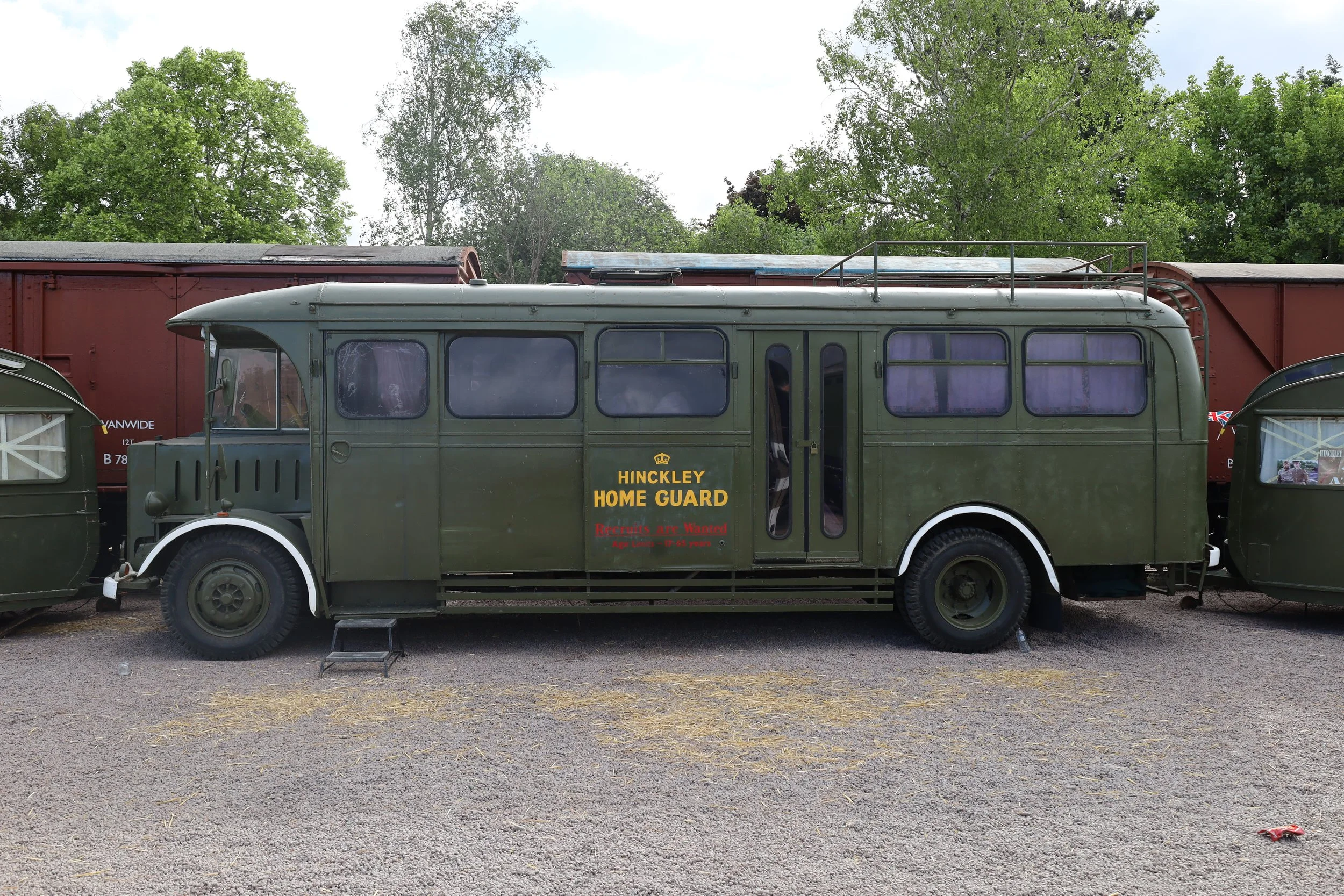 A green vintage bus labeled "Hinckley Home Guard" parked on a gravel surface with train cars and lush green trees in the background.