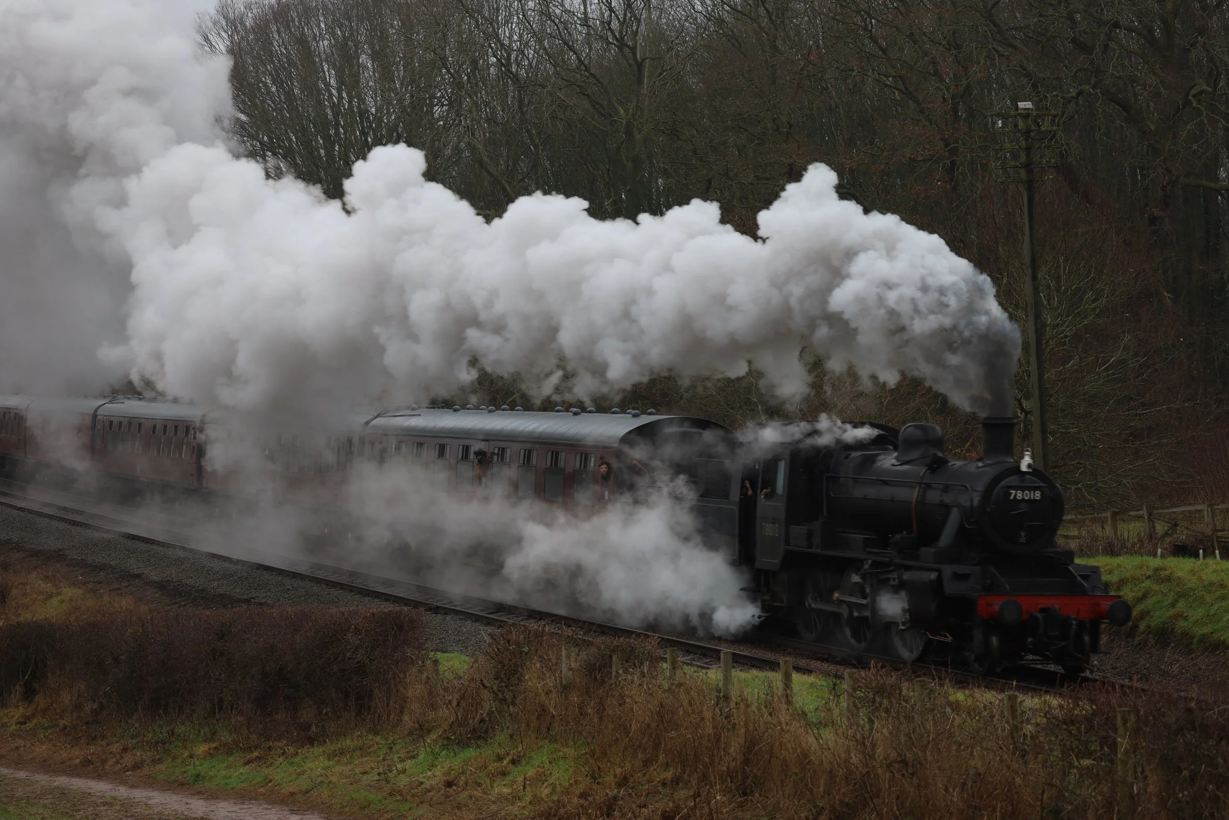 A black steam locomotive pulling passenger cars on a railway track with large white smoke billowing from its chimney, set against a background of leafless trees in a rural area.