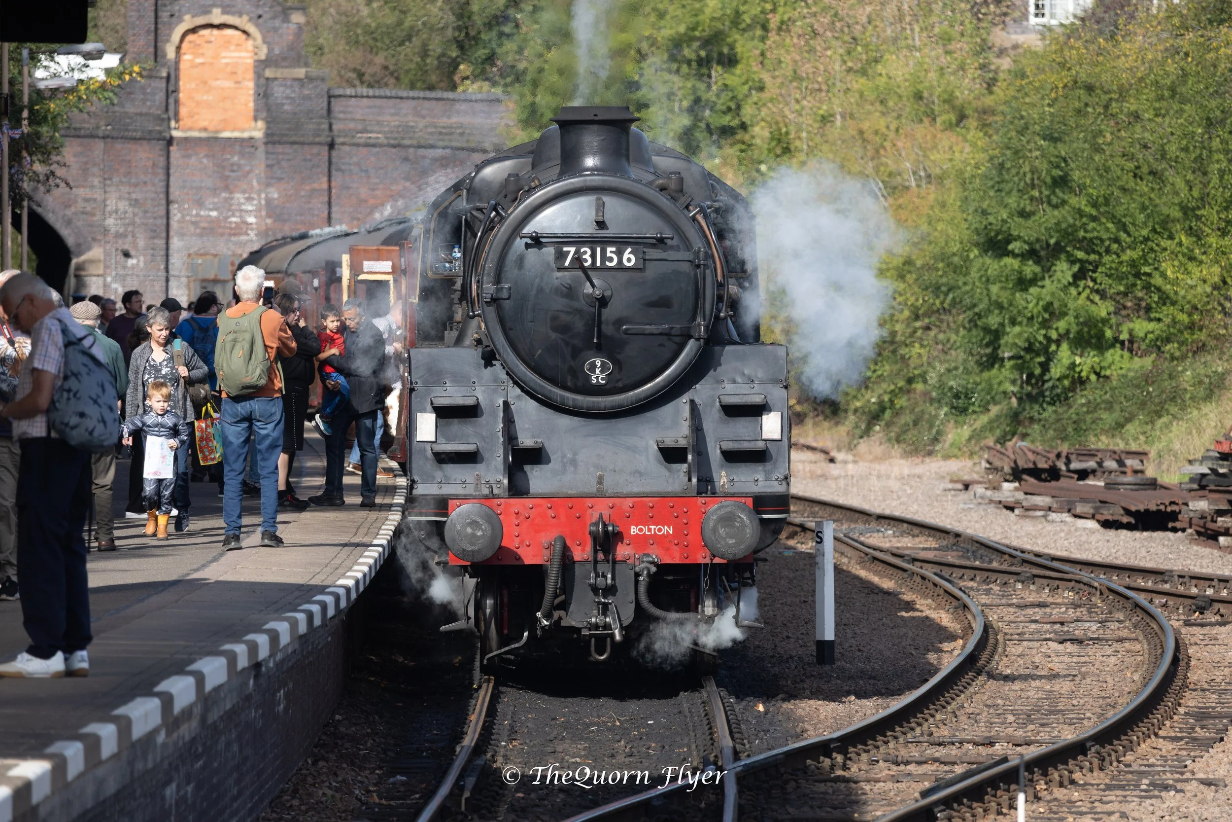 A vintage steam train arriving at a station platform with many people waiting and taking photos. The train is black with red accents and the number 73156 on the front. There are trees in the background and sides of the station visible.
