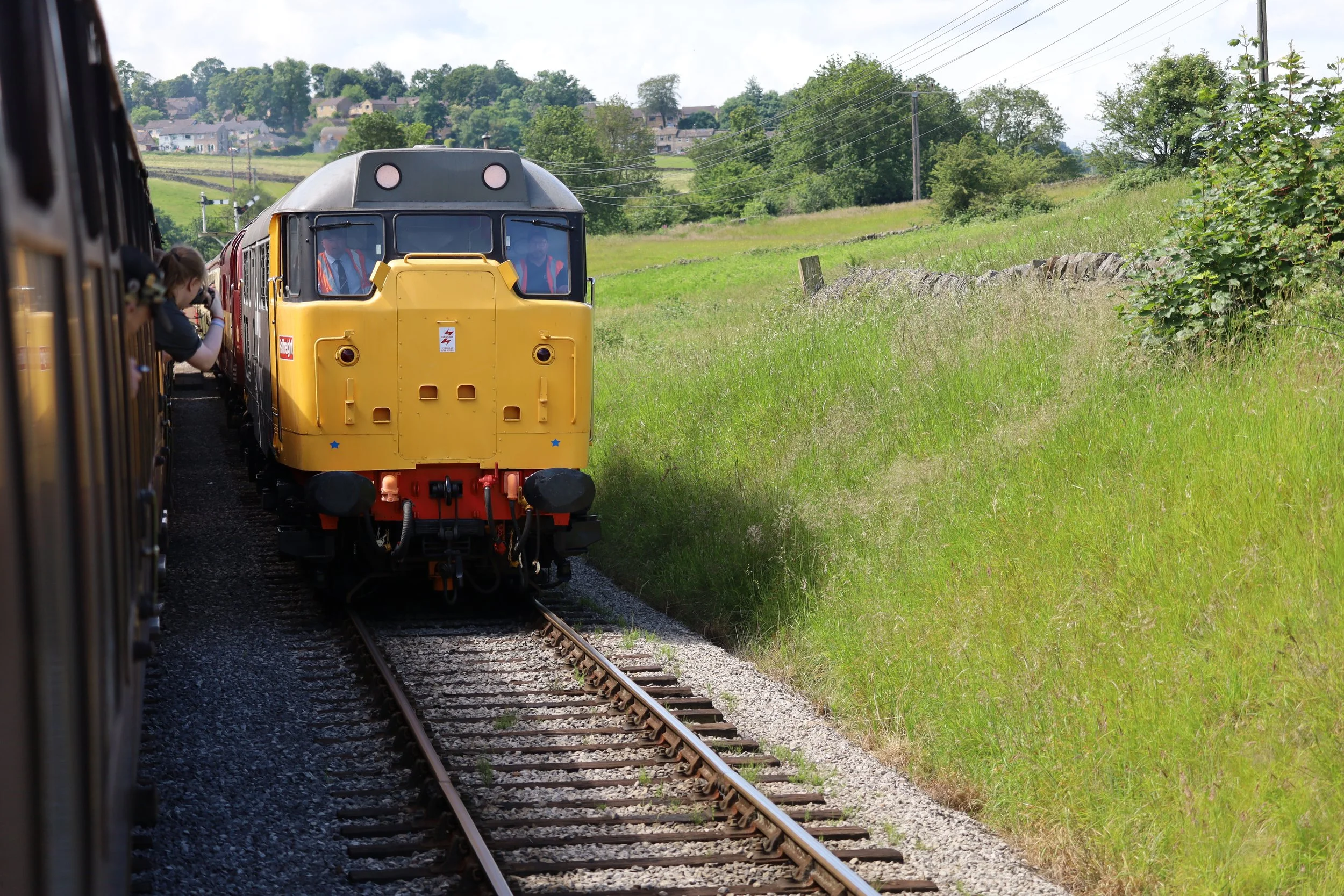 A yellow train is traveling on railroad tracks through a scenic green landscape with trees and hills in the background.