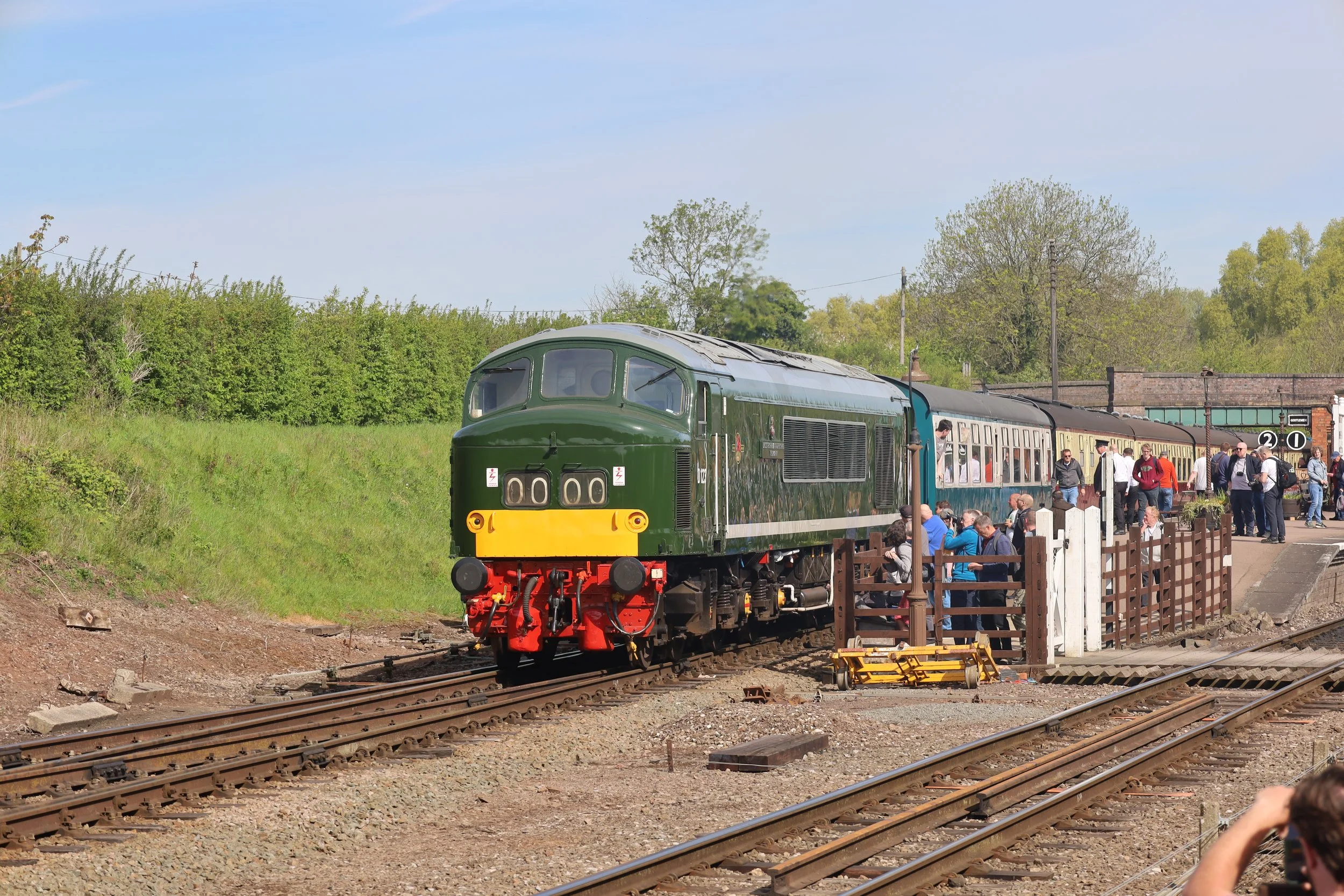 A vintage green and yellow train with a crowd of people boarding and waiting at the station on a sunny day with trees and a blue sky in the background.