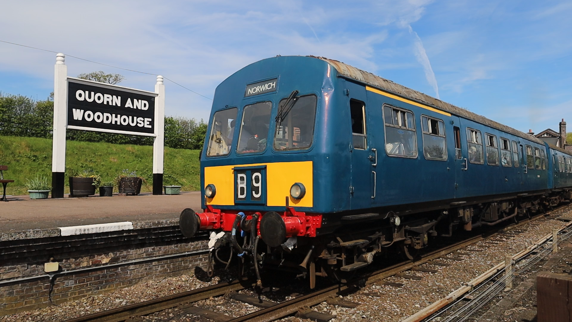 An old blue train with yellow front and red undercarriage, stopped at Quorn and Woodhouse station with a signboard, green grassy hill, and blue sky in the background.