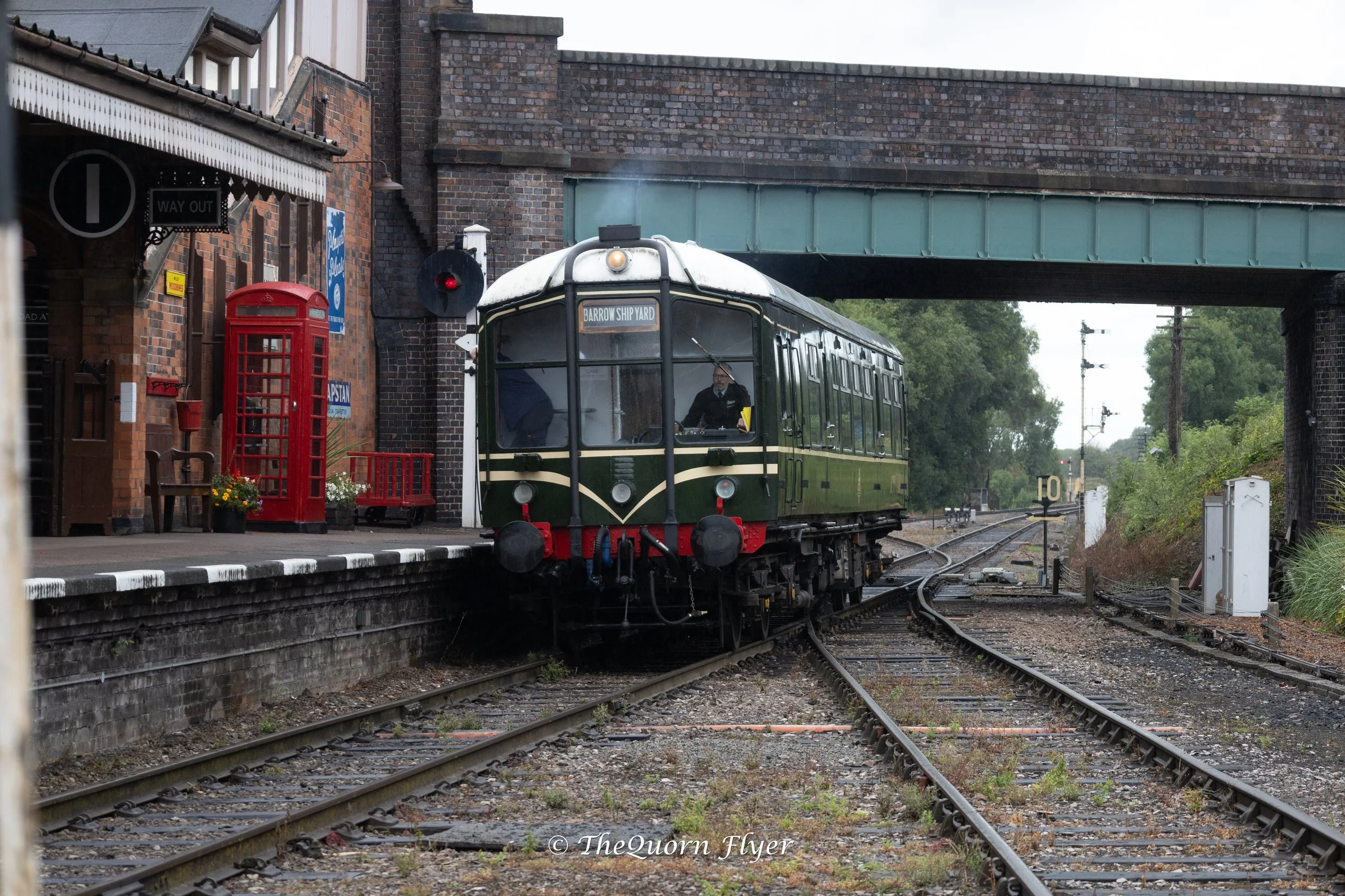 A vintage green train with a black roof and front, moving through a train station under a brick bridge. The station has a brick wall with vintage signage such as a red telephone booth, a 'Way Out' sign, and a blue sign. There are railway tracks and signals, with lush greenery in the background.