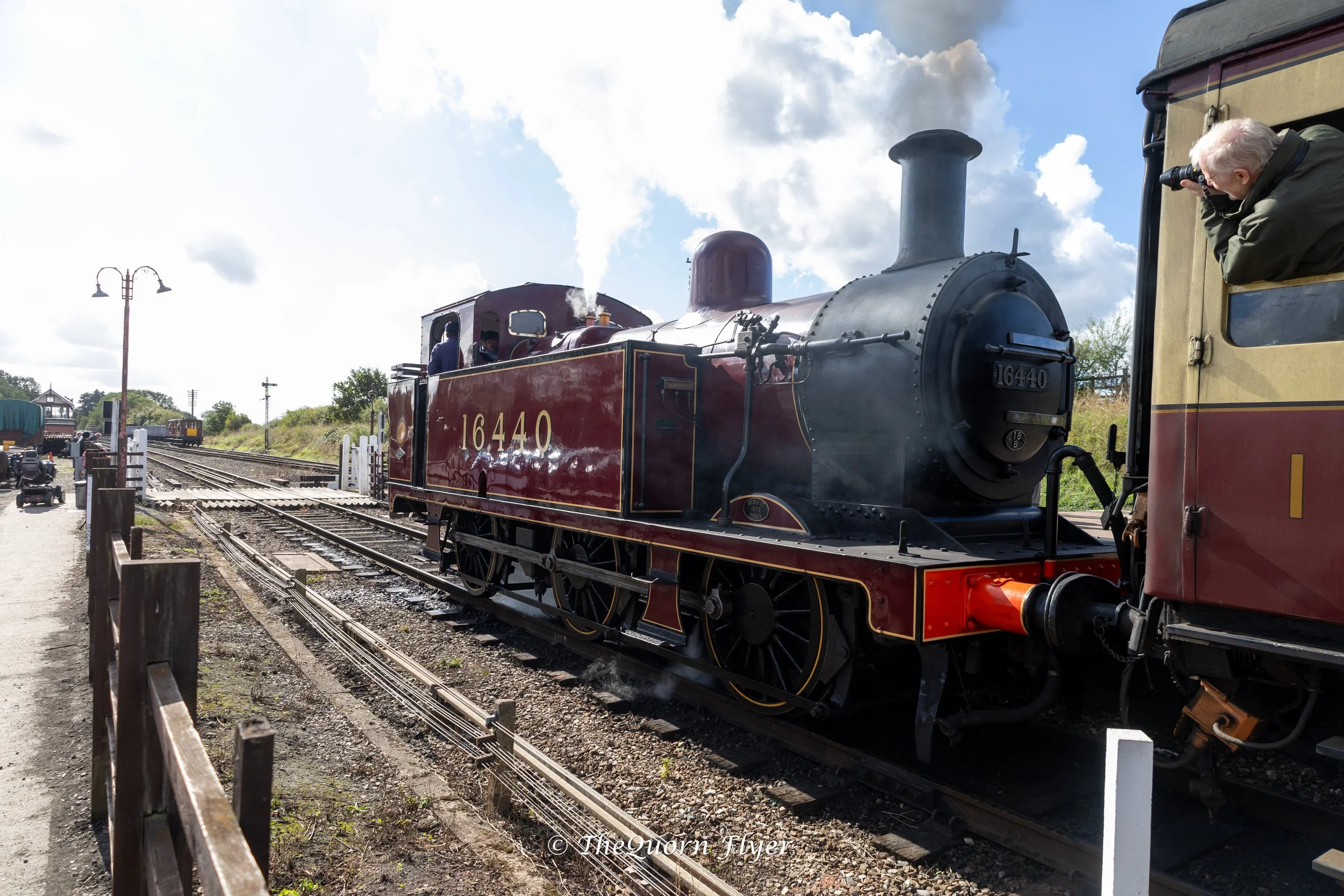 A vintage steam locomotive on train tracks with a man taking a photo, under a partly cloudy sky.