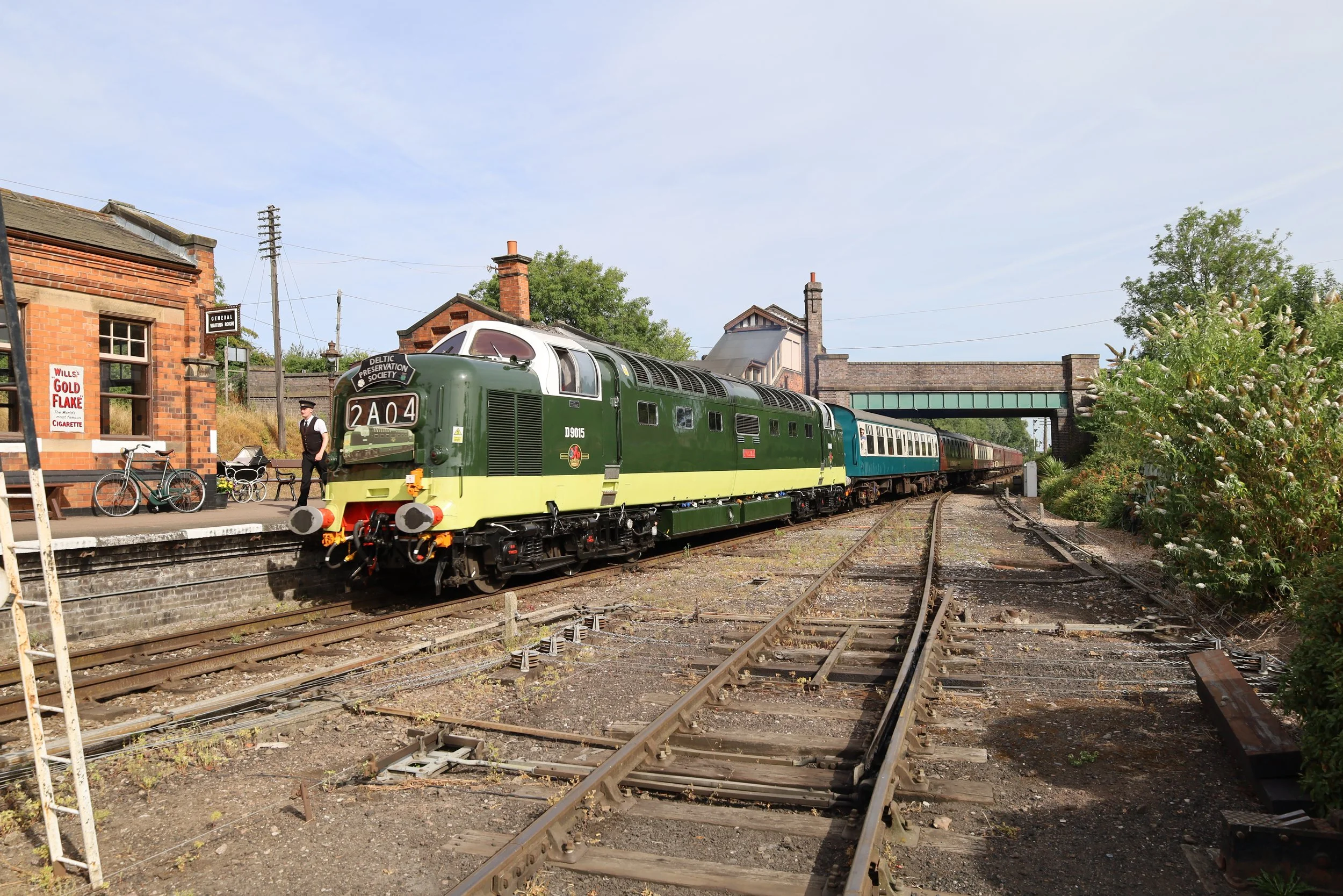 A vintage green train is at a small train station, with a man in vintage attire standing next to it, outside a brick building with a bicycle parked nearby. The train is on old tracks with rusted rails and surrounded by greenery and trees.