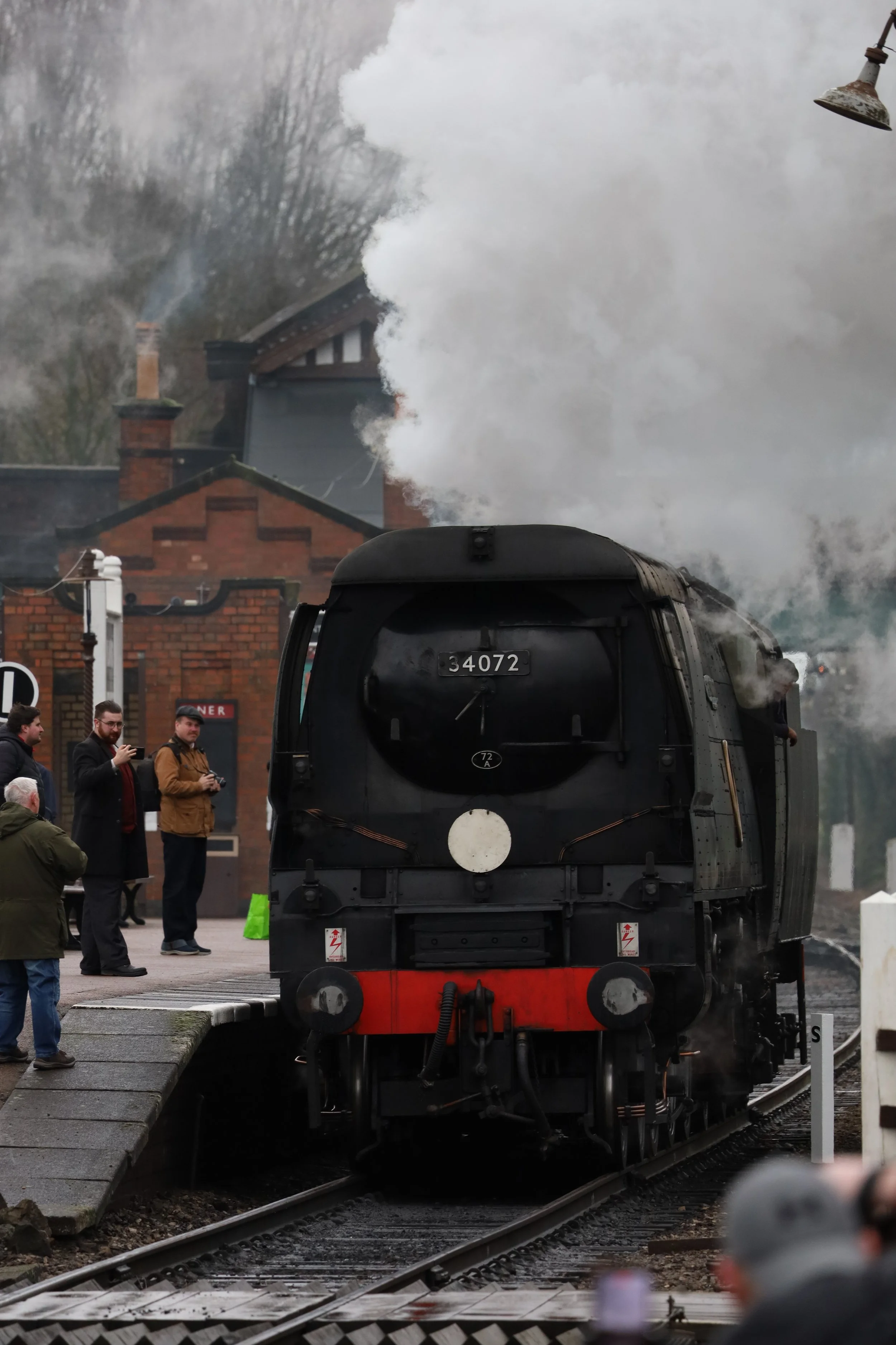 A black steam locomotive with the number 34072 is on railway tracks, emitting a large plume of white steam, with people observing nearby.