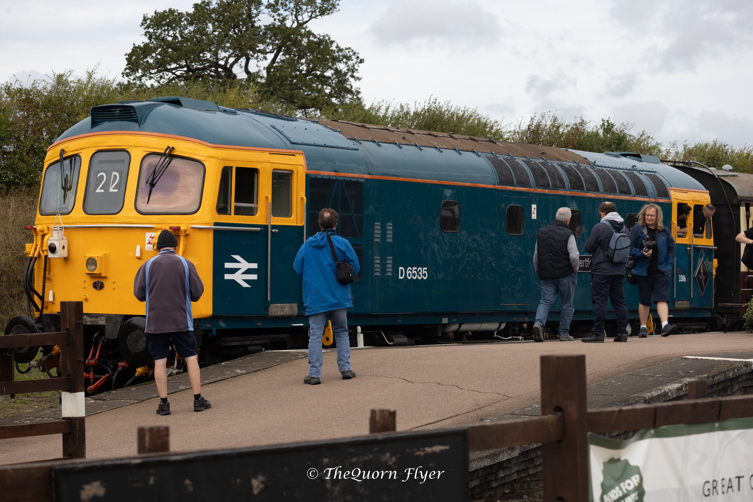 A vintage blue and yellow train locomotive with people standing around it outdoors, some taking photos, under a partly cloudy sky.