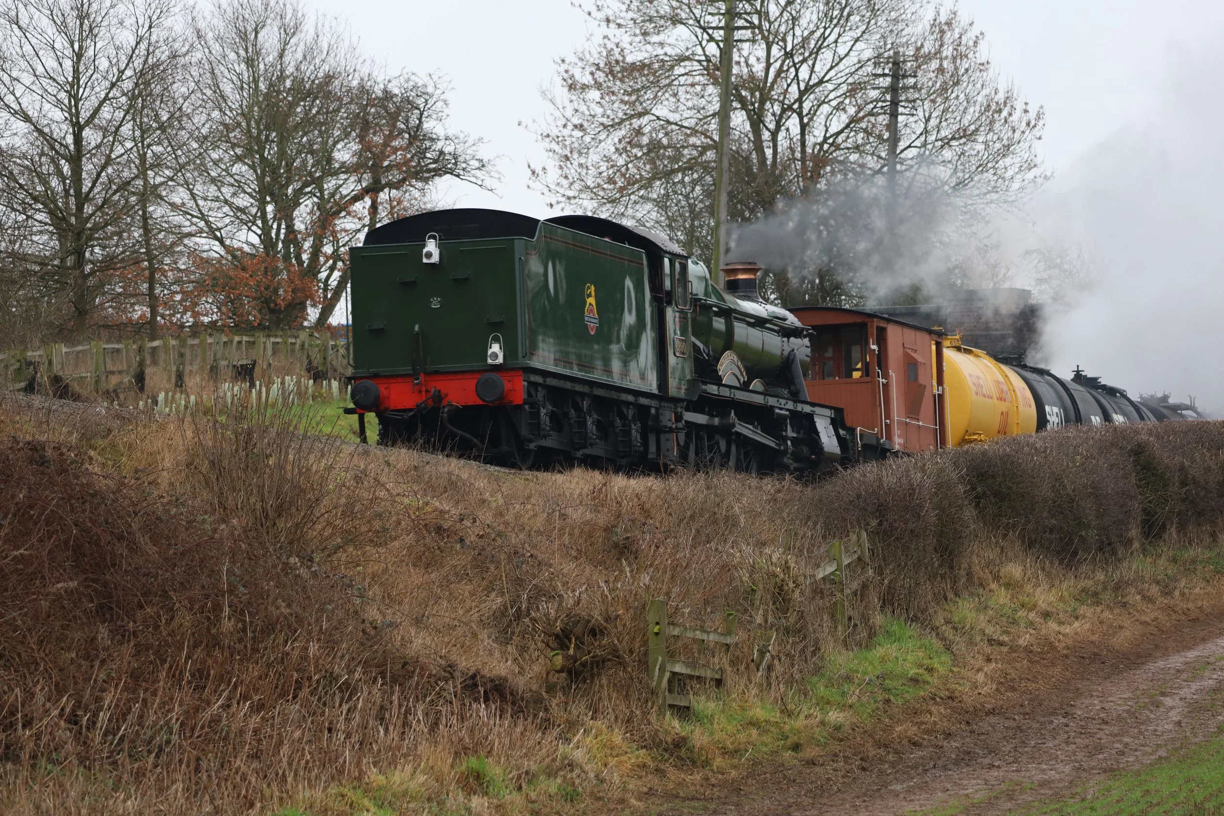 A vintage steam locomotive train with a green engine and yellow tank cars, emitting steam, traveling through a rural area with trees and a wooden fence.