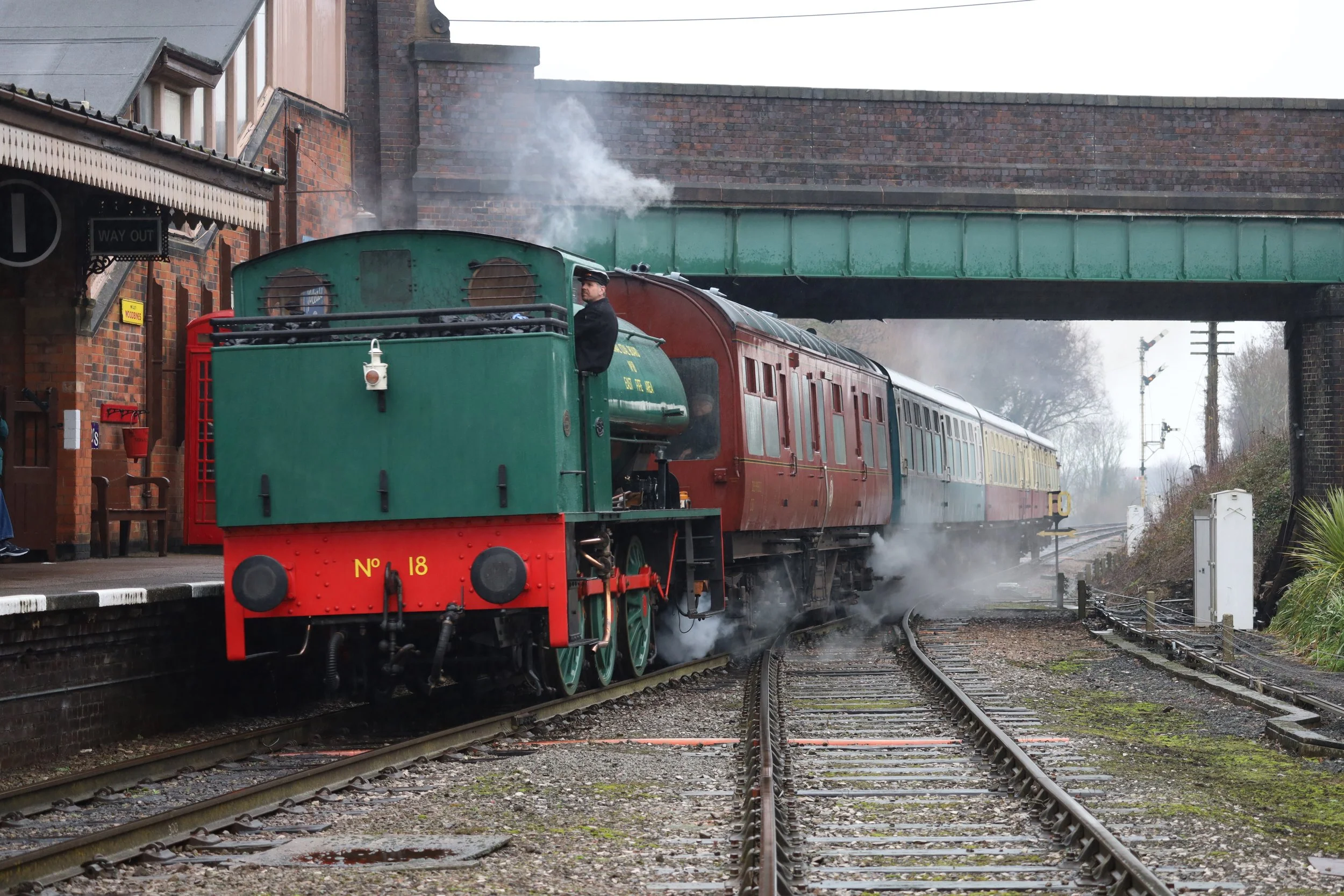 A vintage steam train with a green engine and red front, numbered 18, moving along railway tracks near a brick station building, with smoke emitting from the engine.