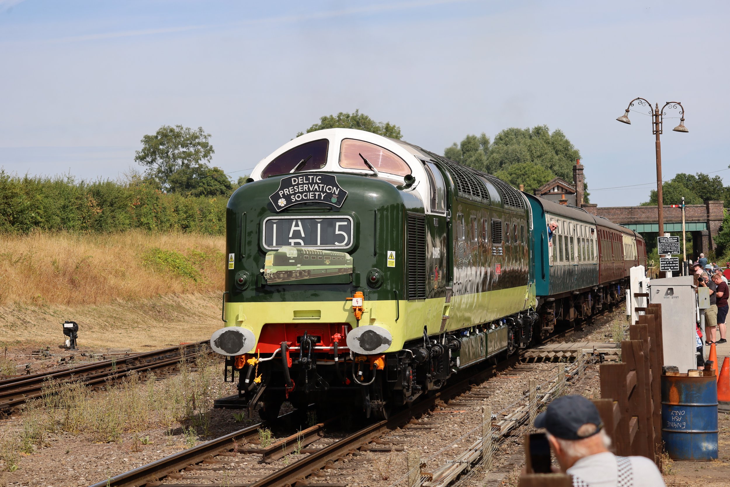 A vintage train painted in green and yellow on tracks during daytime, with people nearby and trees in the background.