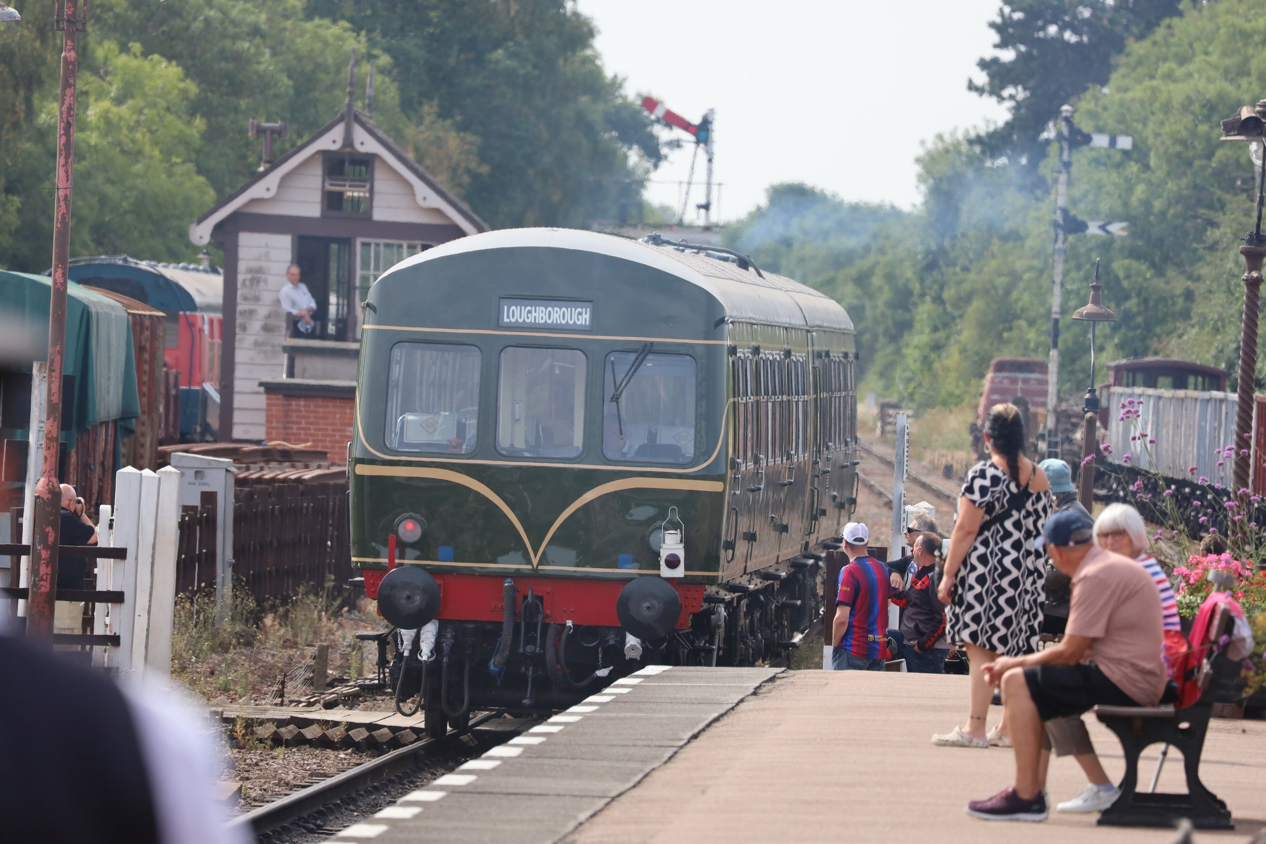 A vintage train labeled 'Loughborough' passing through a rural station with people waiting on the platform. Some are standing, others are sitting on benches, and a woman with a patterned top and glasses is visible in the foreground. The scene includes trees, old station lamps, and a small building in the background.