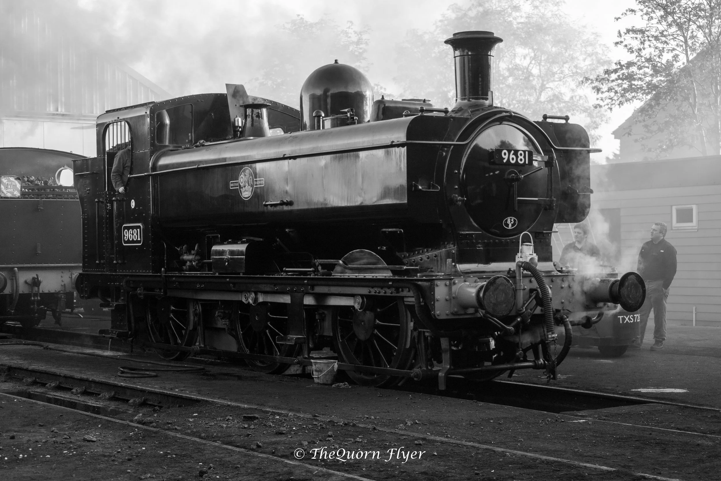A vintage steam locomotive on train tracks with smoke coming from its chimney. Two people are standing nearby, and a building is visible in the background. The photo is black and white.
