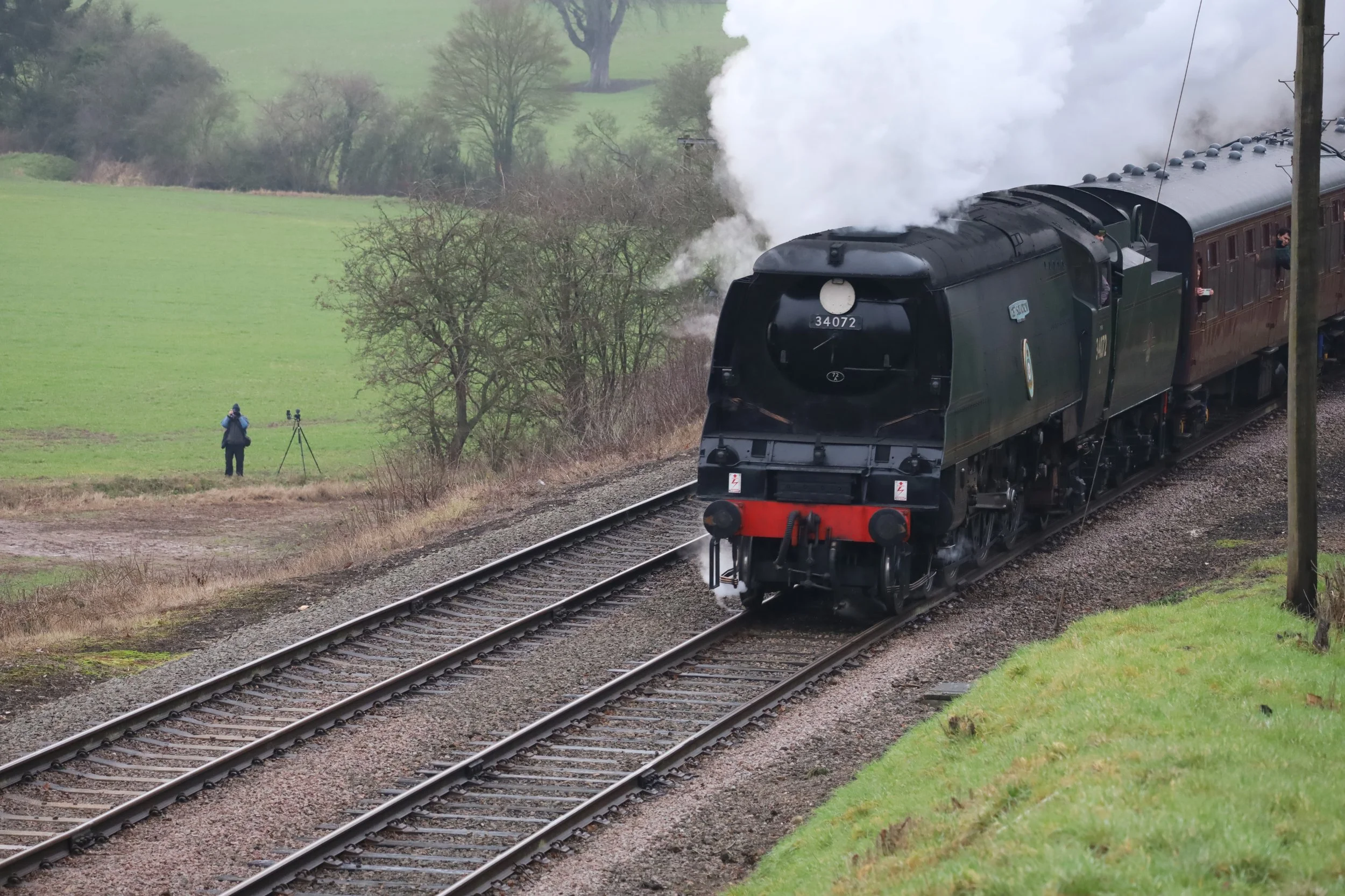 A black steam locomotive train traveling on tracks through a rural area with green fields and trees, with smoke billowing from the engine, and a person standing nearby with a camera on a tripod.
