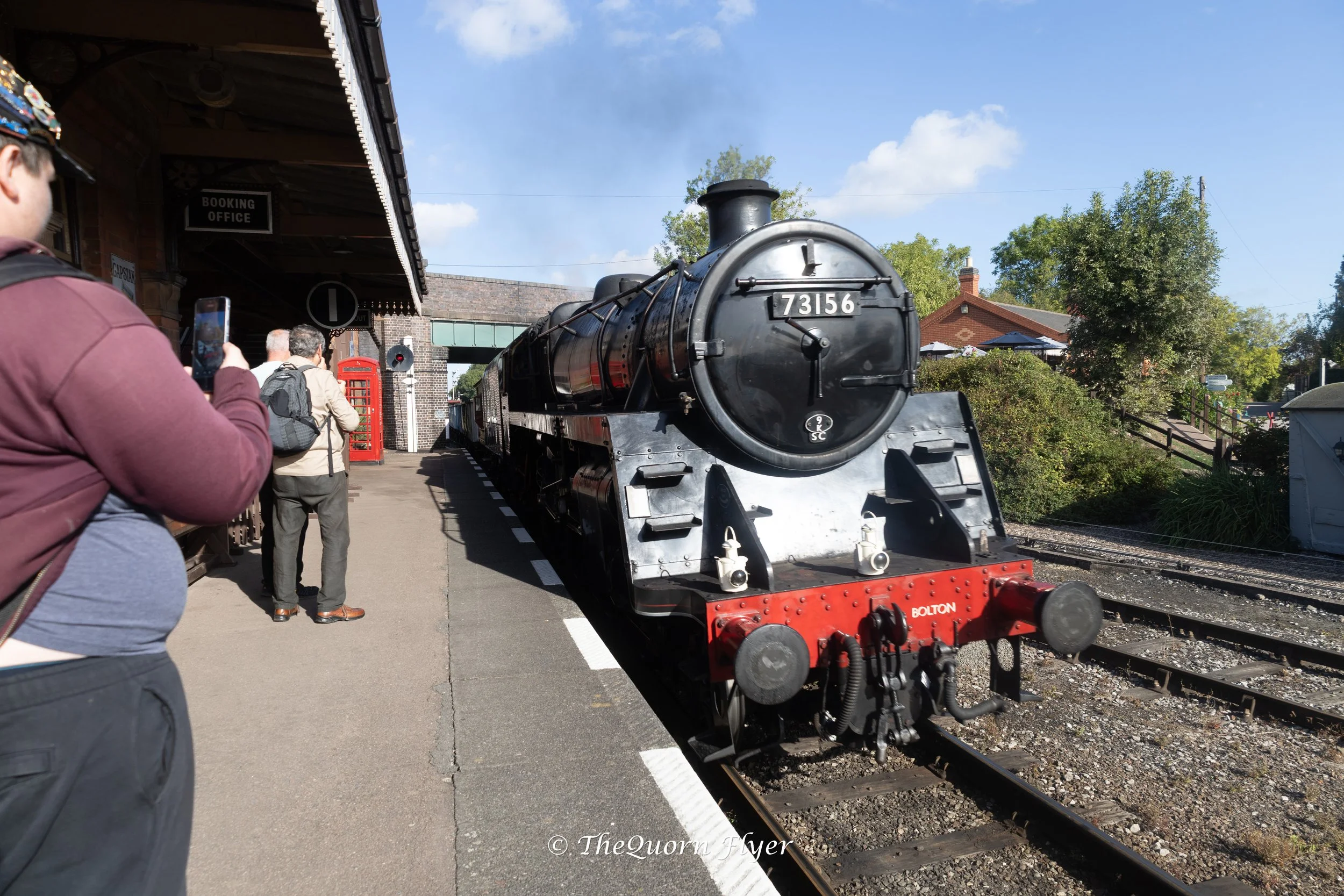 A black steam locomotive train with the number 73156 is on a train track at a station, with people waiting nearby and one person taking a photo. The station building has a sign for the booking office and queued people are visible. Trees and a blue sky with some clouds are in the background.
