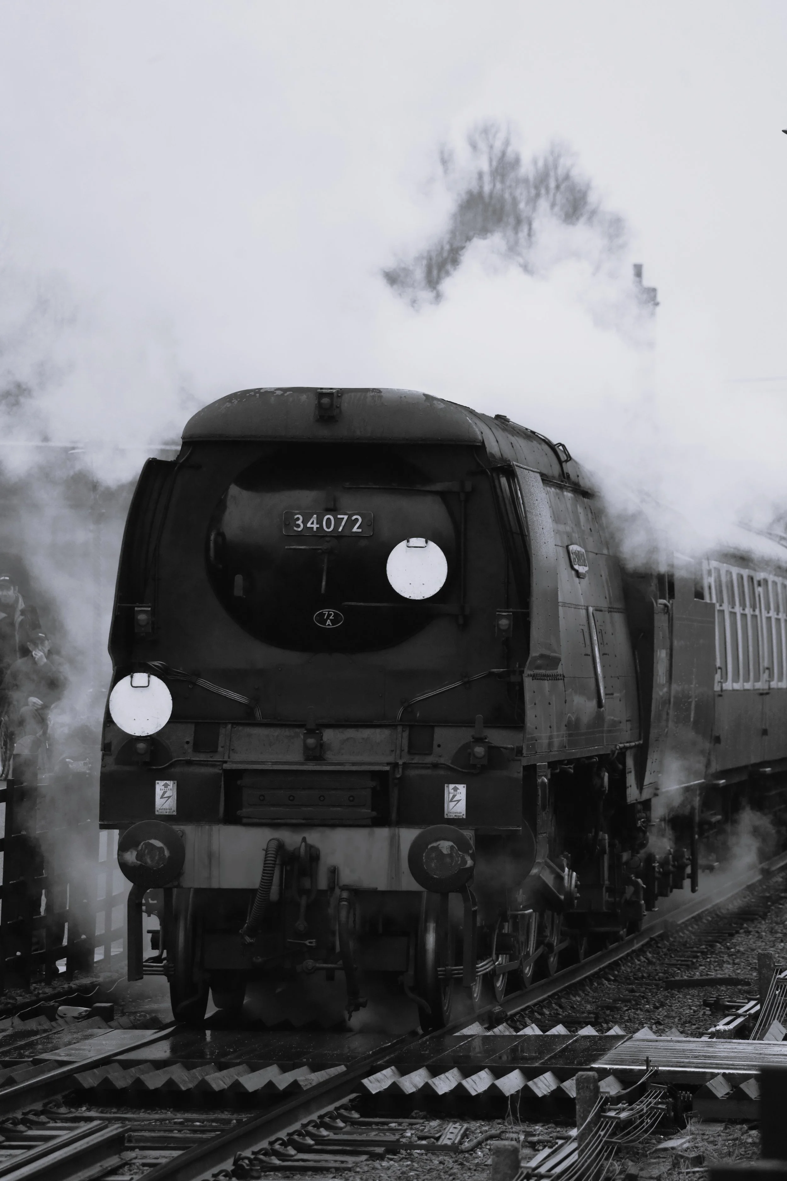 A black steam locomotive train on the tracks emitting steam, with trees and railway infrastructure visible in the background.
