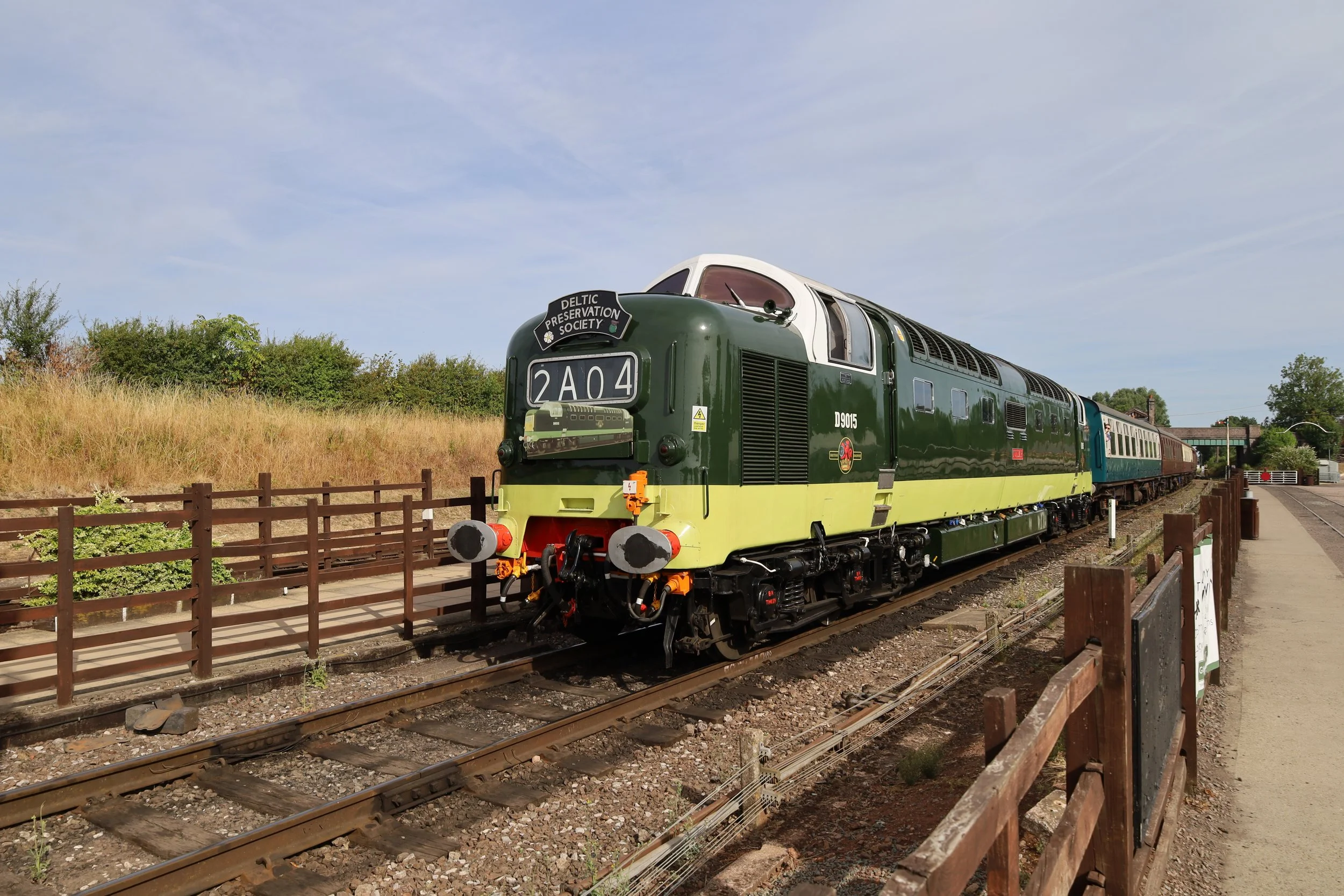 Vintage green and yellow train with the sign 'Deltic Preservation Society', parked on train tracks next to a walkway and wooden fence, under a partly cloudy sky.