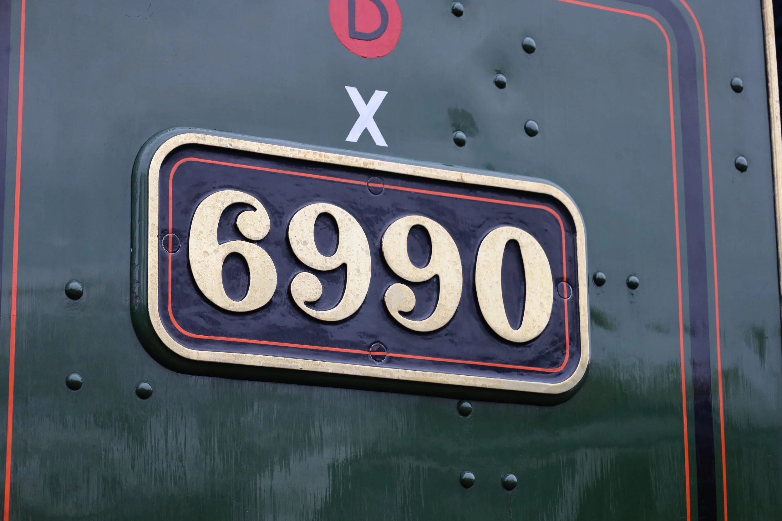 A close-up of a vintage license plate with the number 6990, mounted on a dark green surface with visible rivets and red and orange striping.