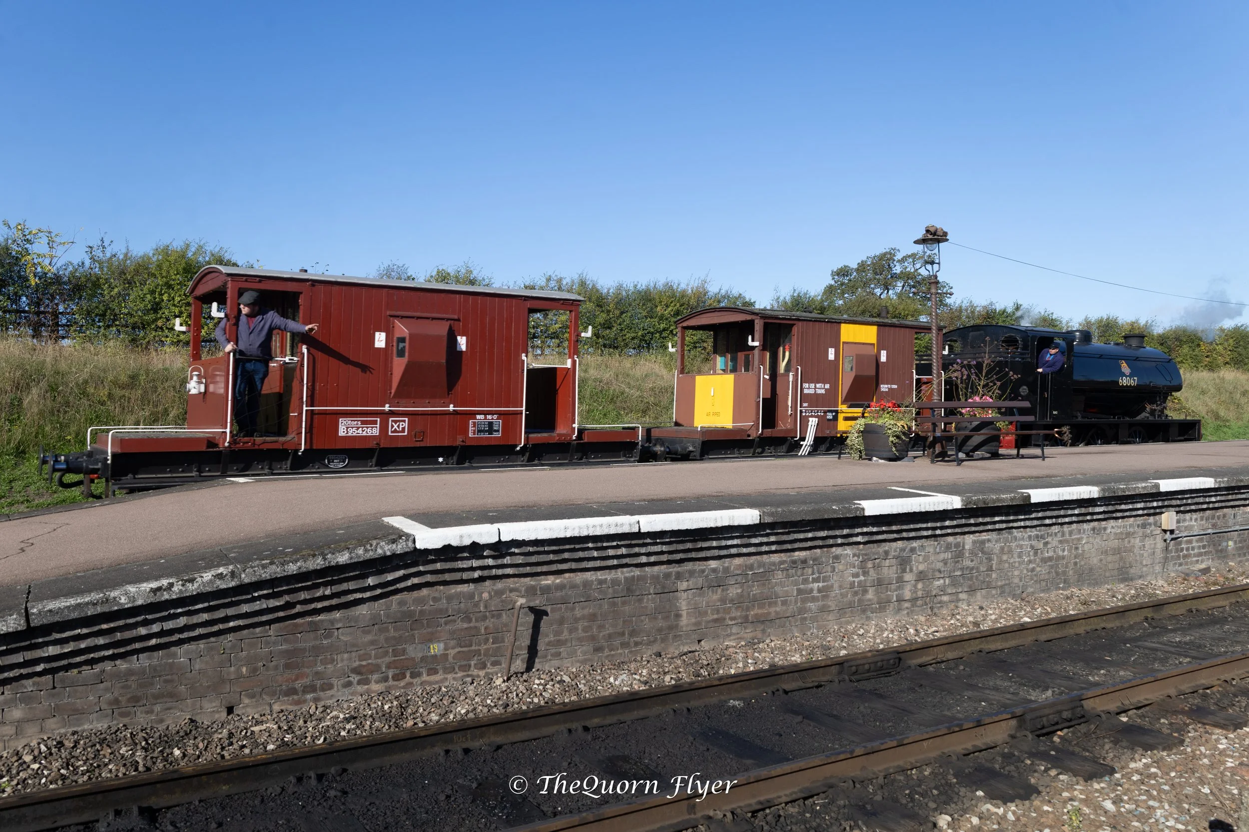 A small vintage train with black engine and red carriages at a railway station or trackside, with a person standing on the first carriage and another near the engine, a telephone pole with a line, and greenery in the background under a clear blue sky.