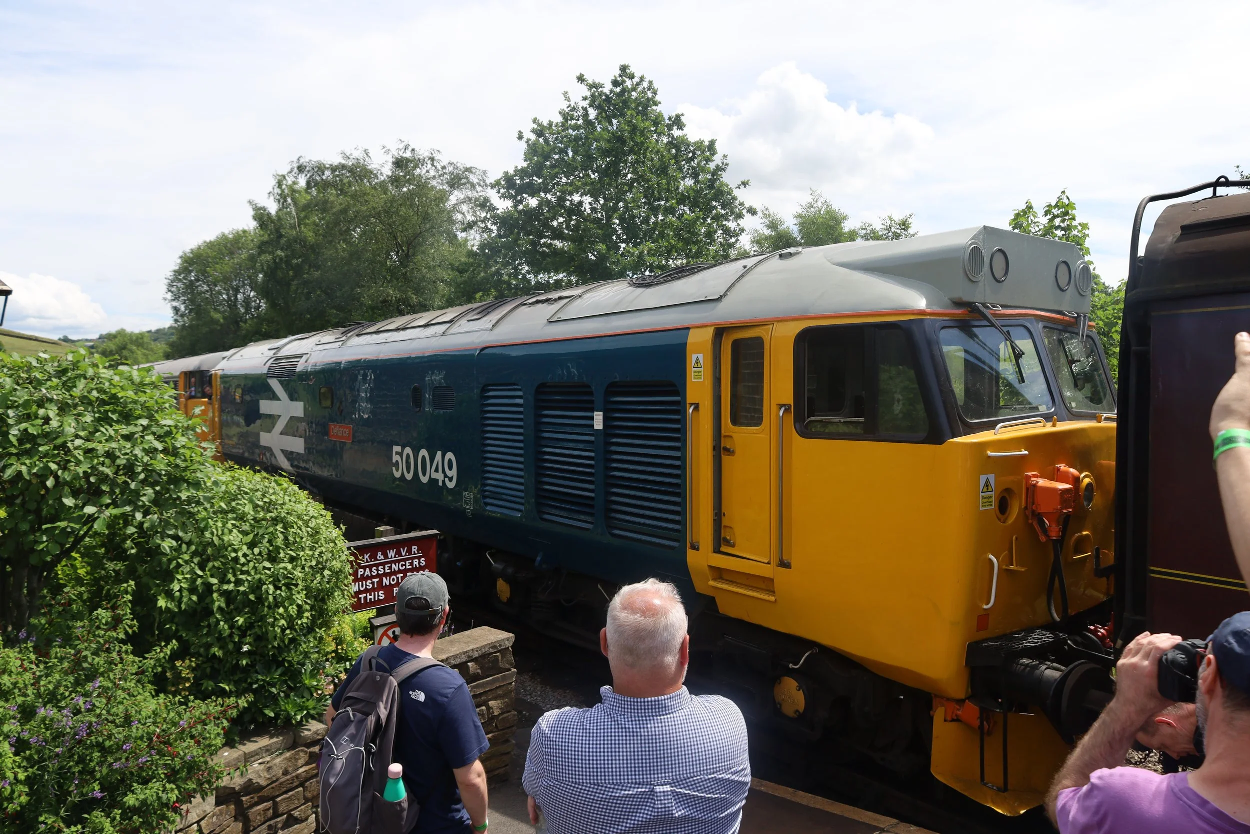 A vintage yellow and blue train with the number 50049 on its side, surrounded by a group of people, some taking photos, outdoors with trees and a partly cloudy sky in the background.