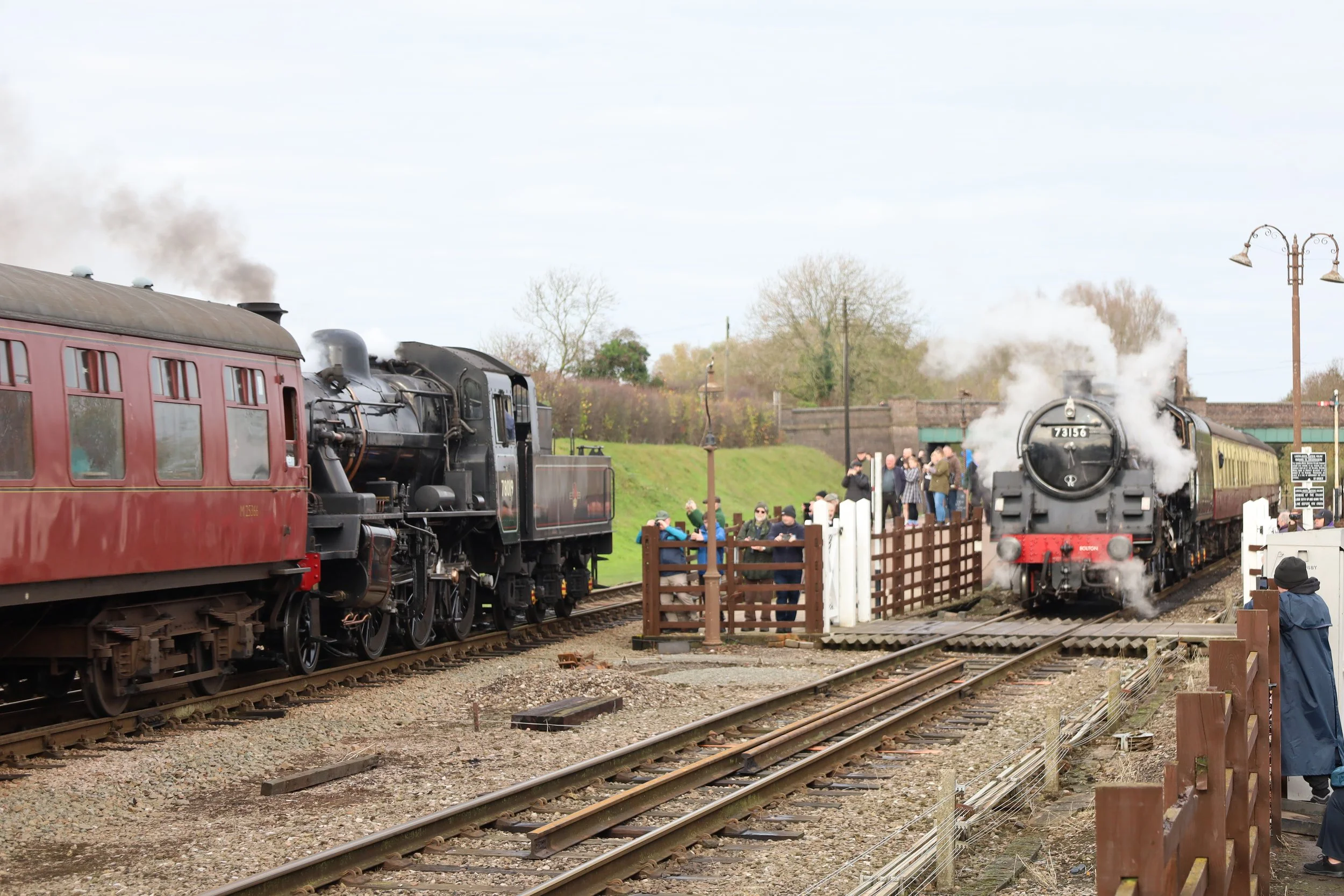 Two vintage steam locomotives on a train track with passengers and onlookers nearby, steam rising from the engines and trees in the background.