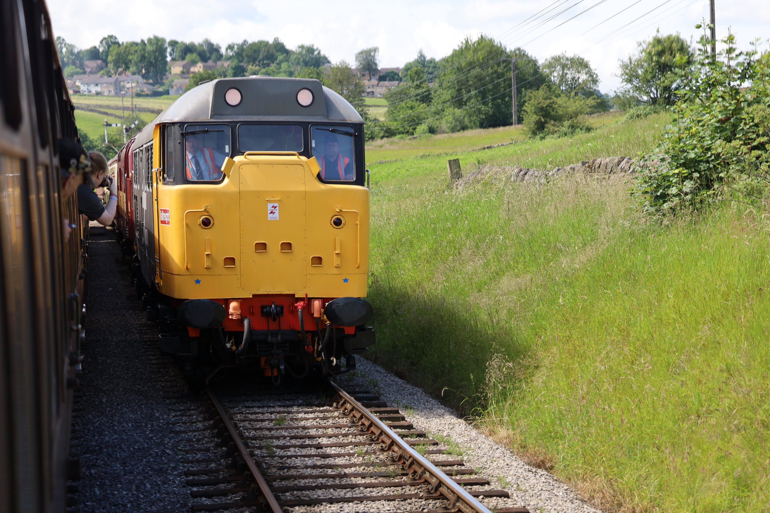 A yellow locomotive train moving along railway tracks through a green countryside with grassy fields and trees, with passengers visible on the left side of the train and houses on a hill in the background.
