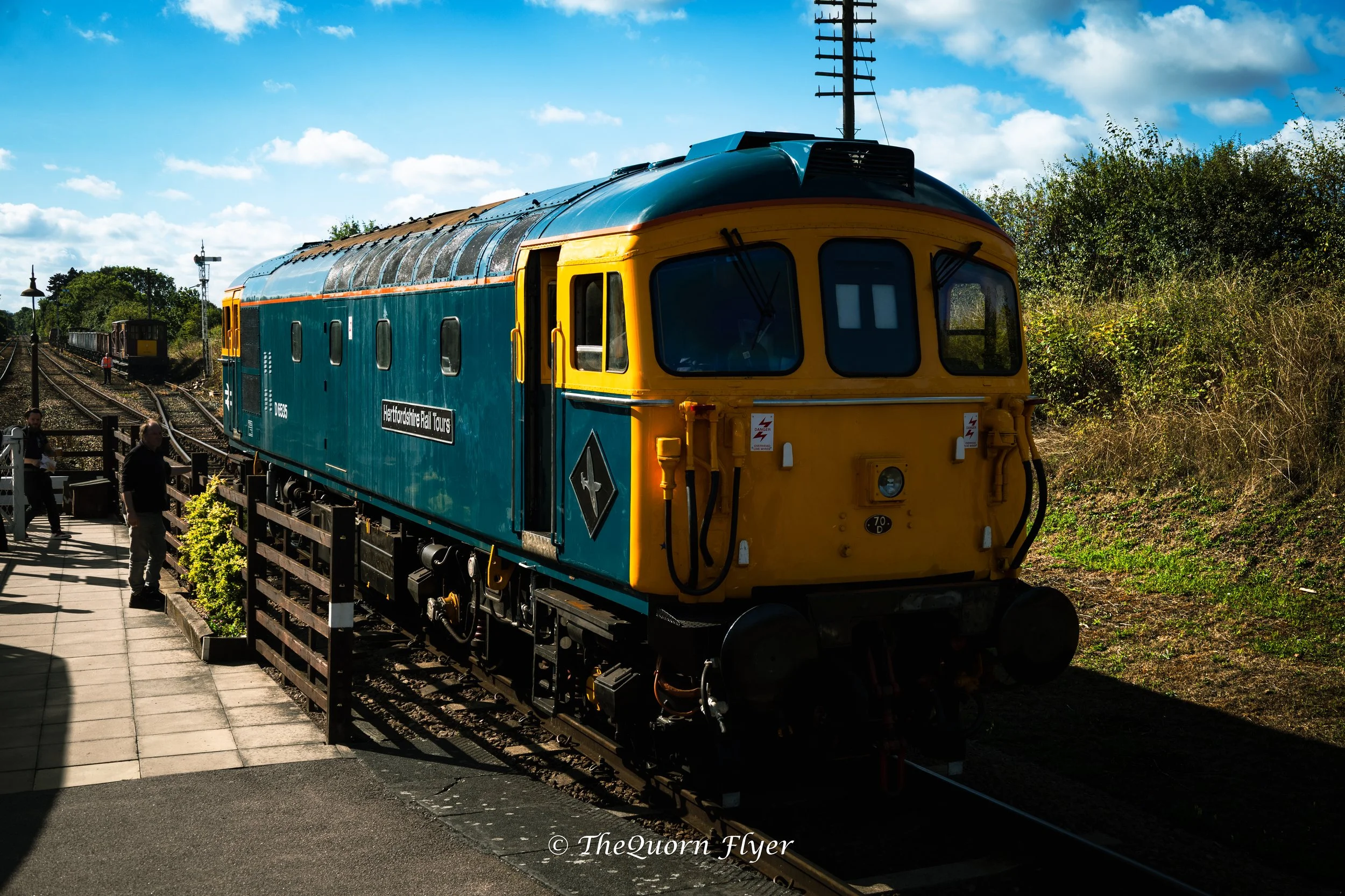 A historic blue and yellow train on a railway track outdoors, with people nearby, under a partly cloudy sky.