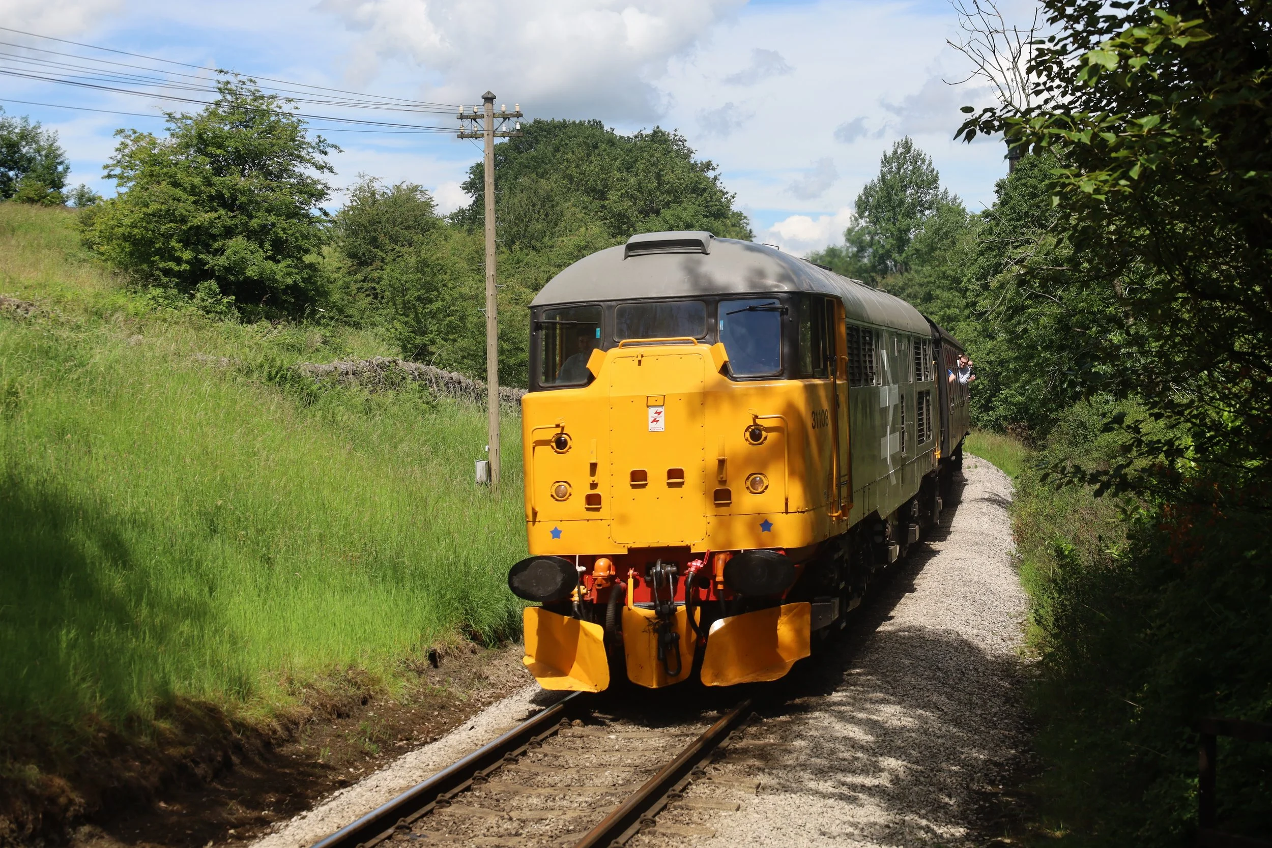 A yellow locomotive train moving along a narrow track through a lush, green landscape with trees and hills under a partly cloudy sky.