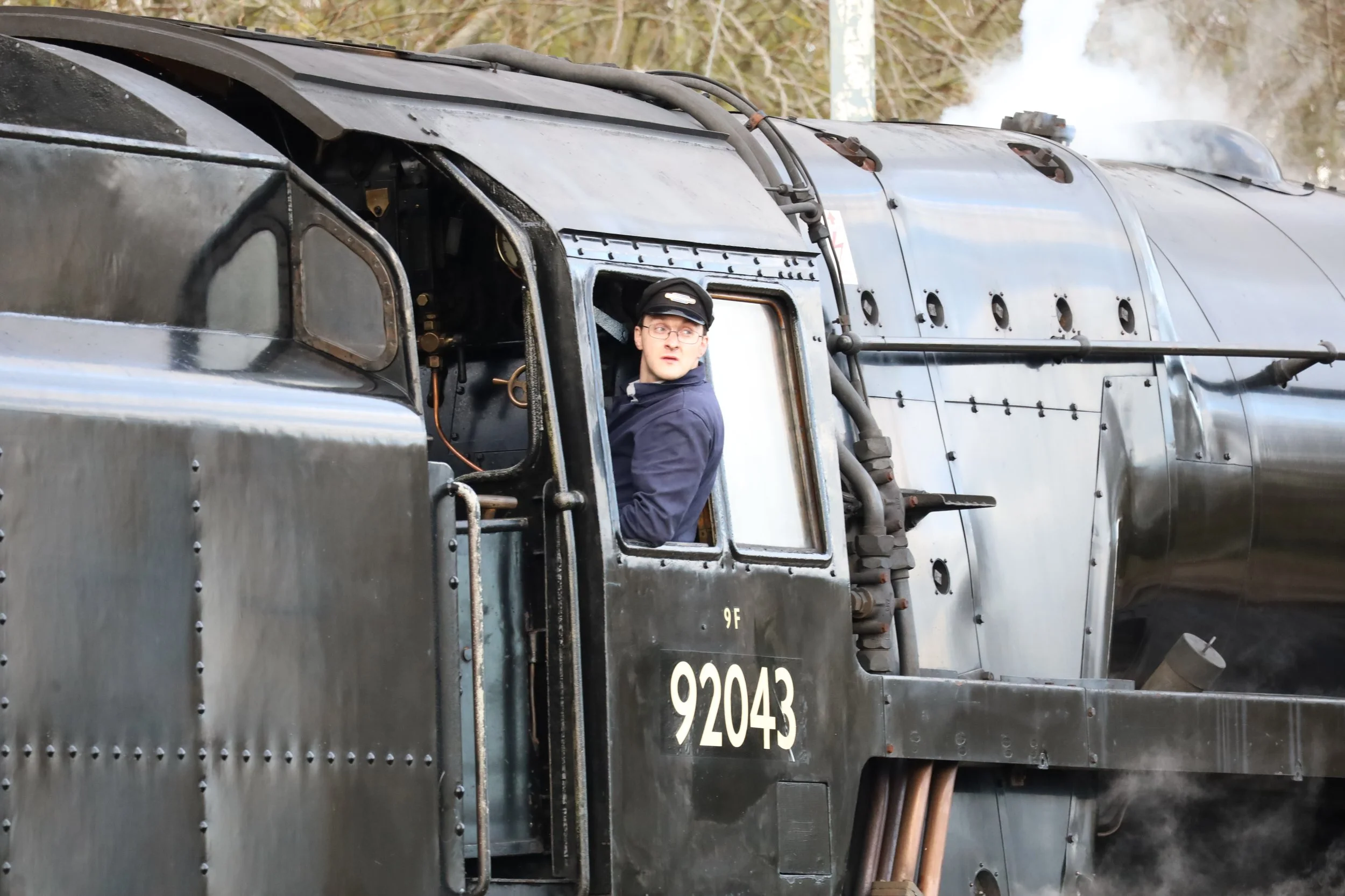 A man with glasses and a black cap is sitting in the cockpit of a vintage military tank with an open hatch, with steam or smoke rising in the background.