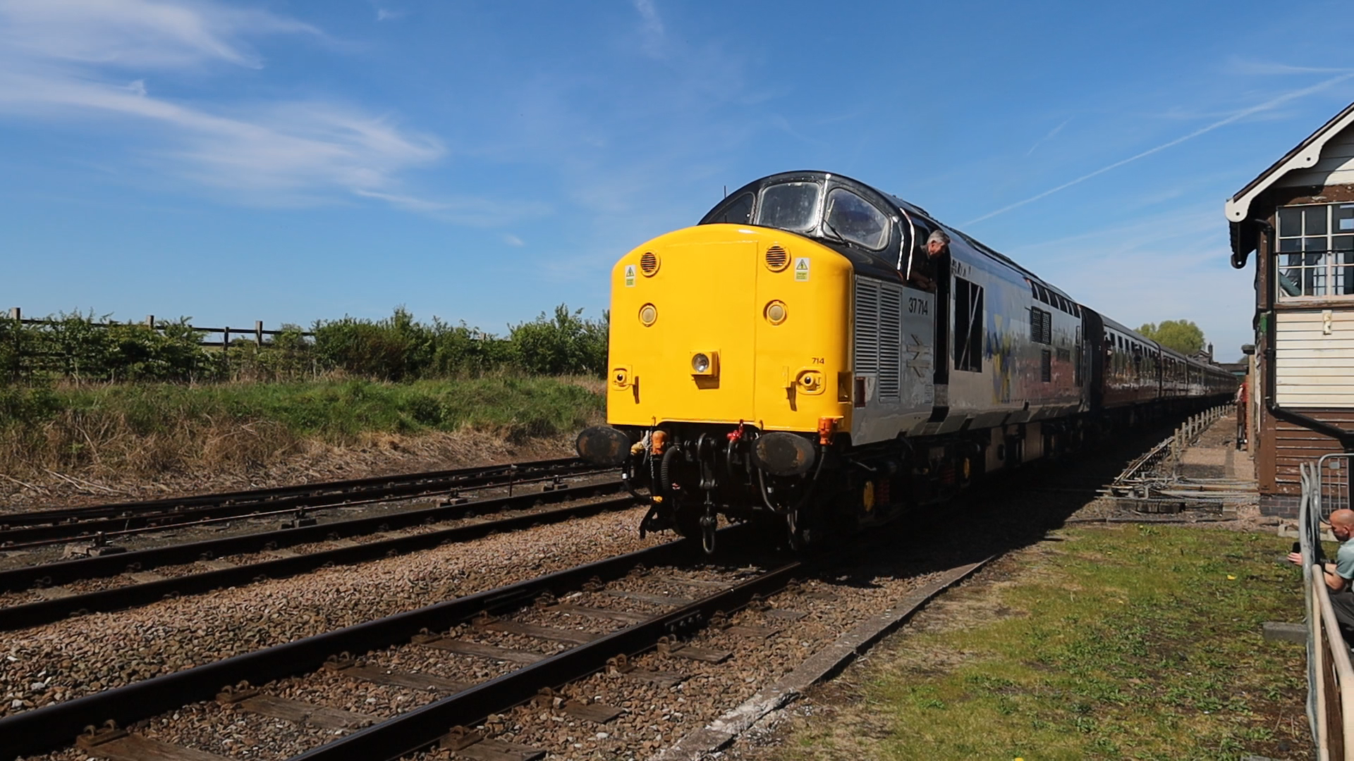 A vintage yellow and black train on railway tracks beside a small station building under a clear blue sky.