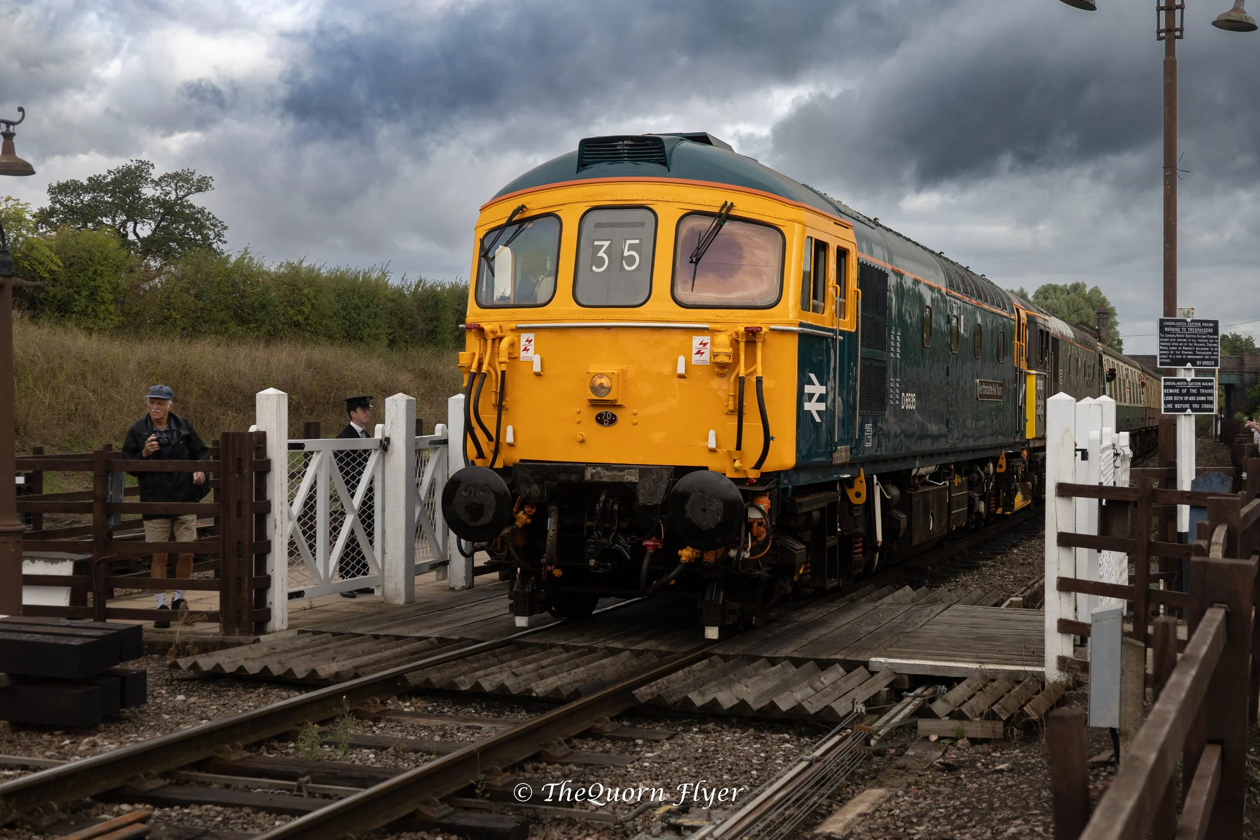 A vintage train with a yellow front and dark green body, marked with the number 35, is at a station with overcast skies. There are two people in vintage attire near a white fence, one holding a camera, and railway tracks with wooden planks in the foreground. A sign warns about train crossing safety.