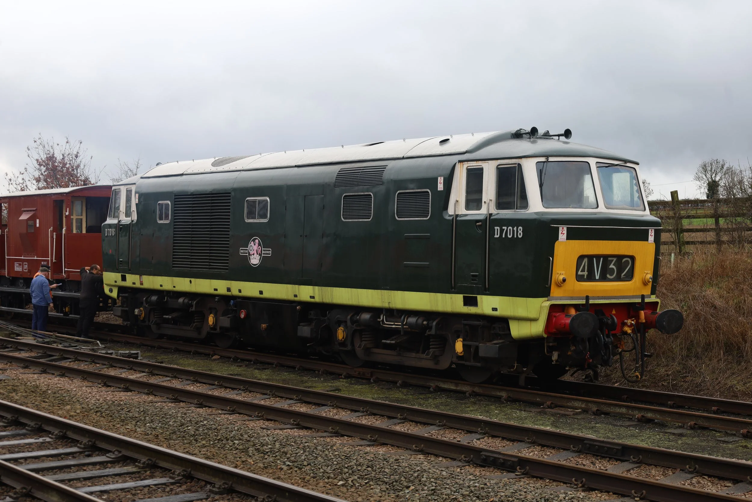 A vintage train with a dark green and yellow locomotive and a red carriage is on railway tracks outdoors with overcast skies and trees in the background.