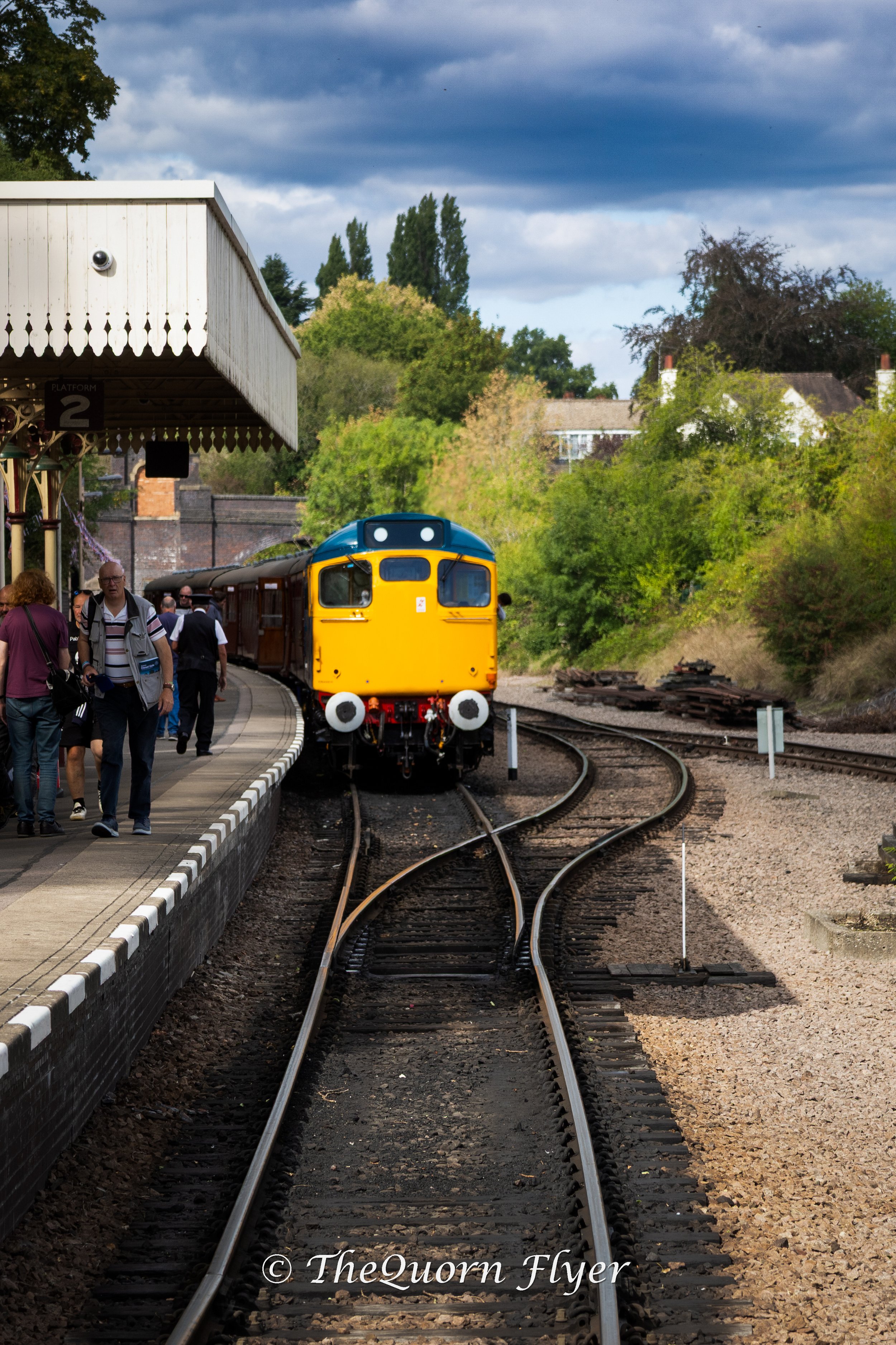 A yellow train approaching a platform at a train station with people walking on the platform and a garden with trees and houses in the background, under a partly cloudy sky.