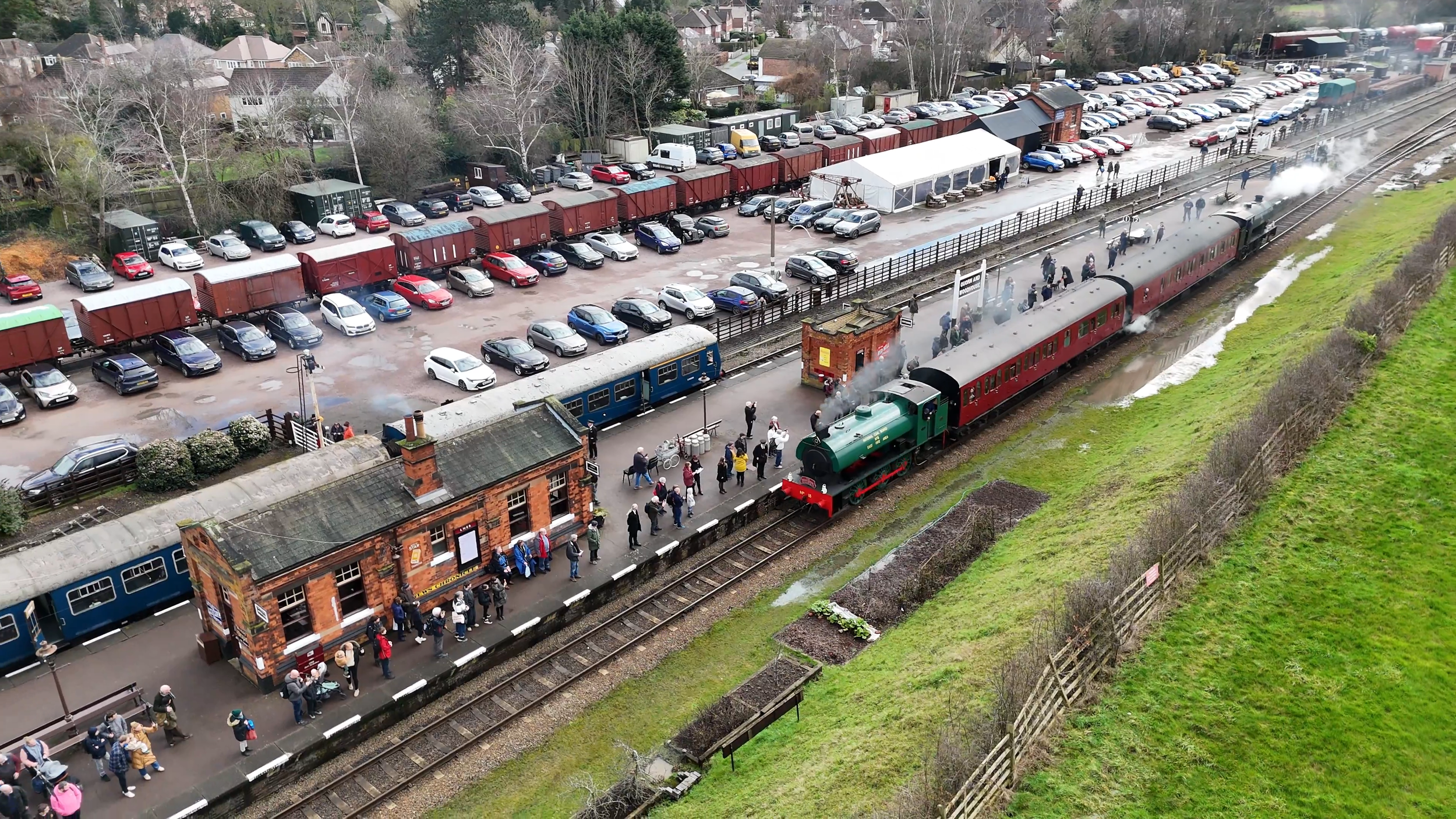 An aerial view of a train station with a steam train arriving or departing, full of passengers on the platform. The platform is lined with parked cars and some containers, while a residential neighborhood with houses and trees is visible in the background.