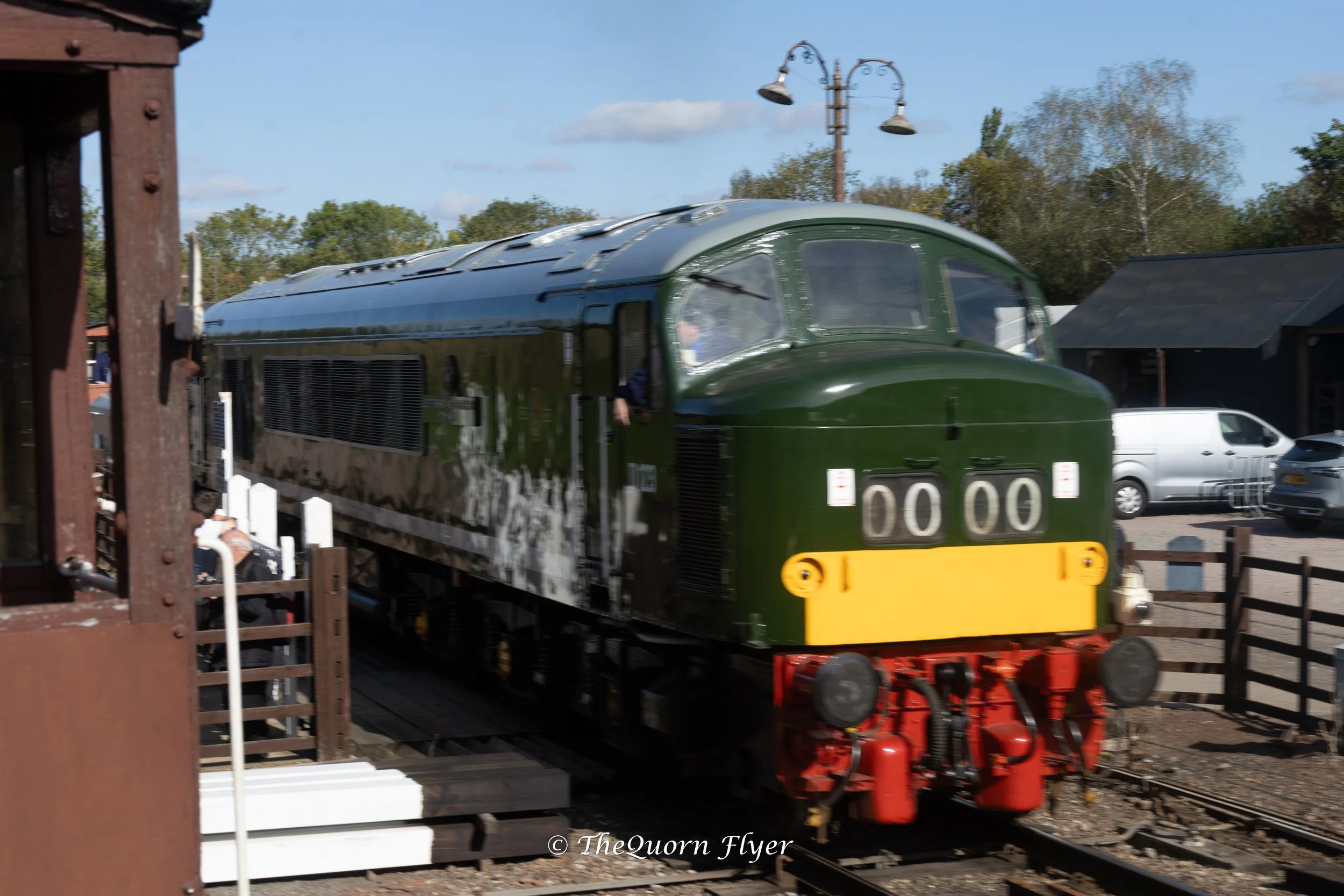 A vintage train painted green and silver moving on a railway track during daytime, with trees and a blue sky in the background.