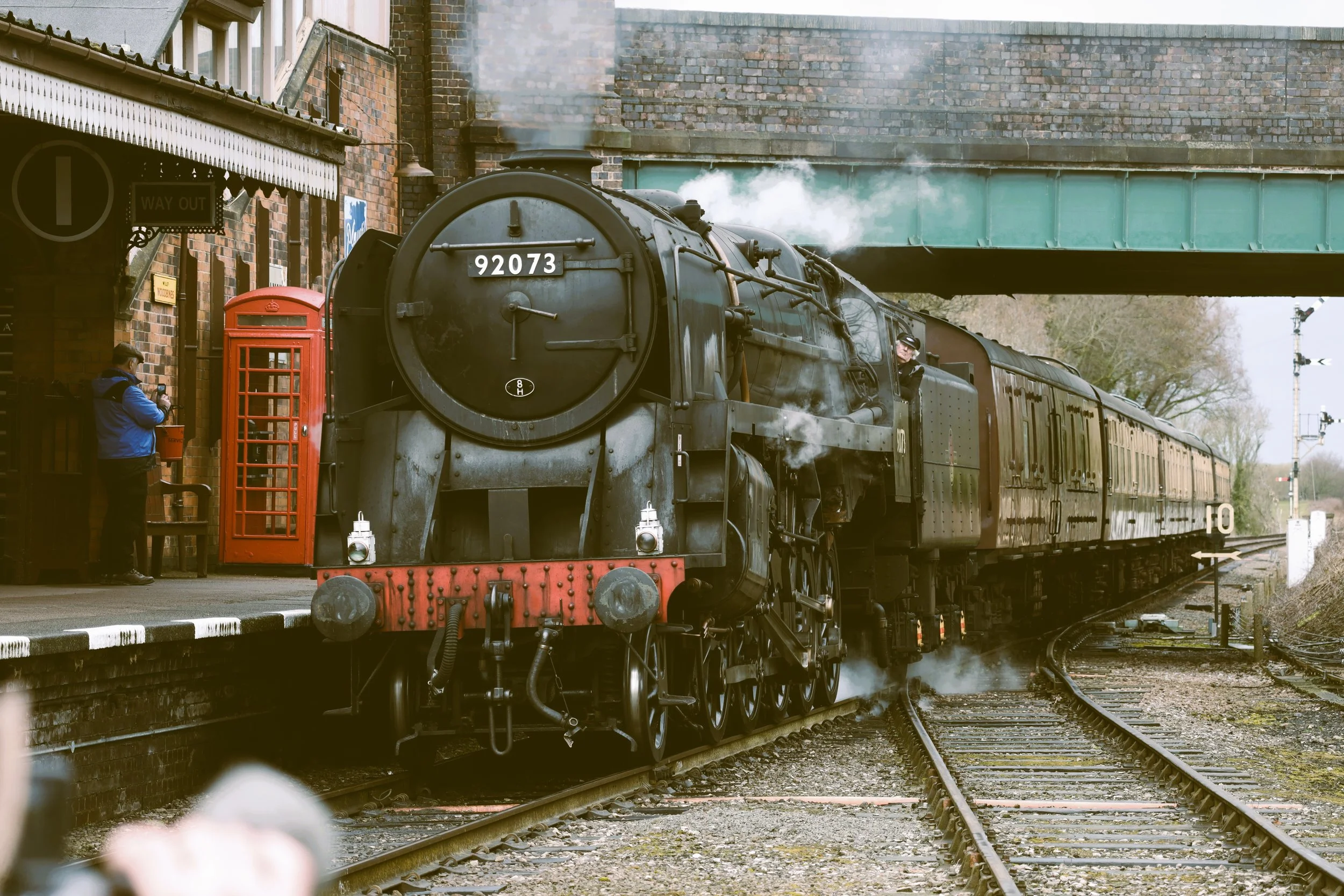 A vintage steam train on the tracks at a station with a person taking photo and a red telephone booth nearby.