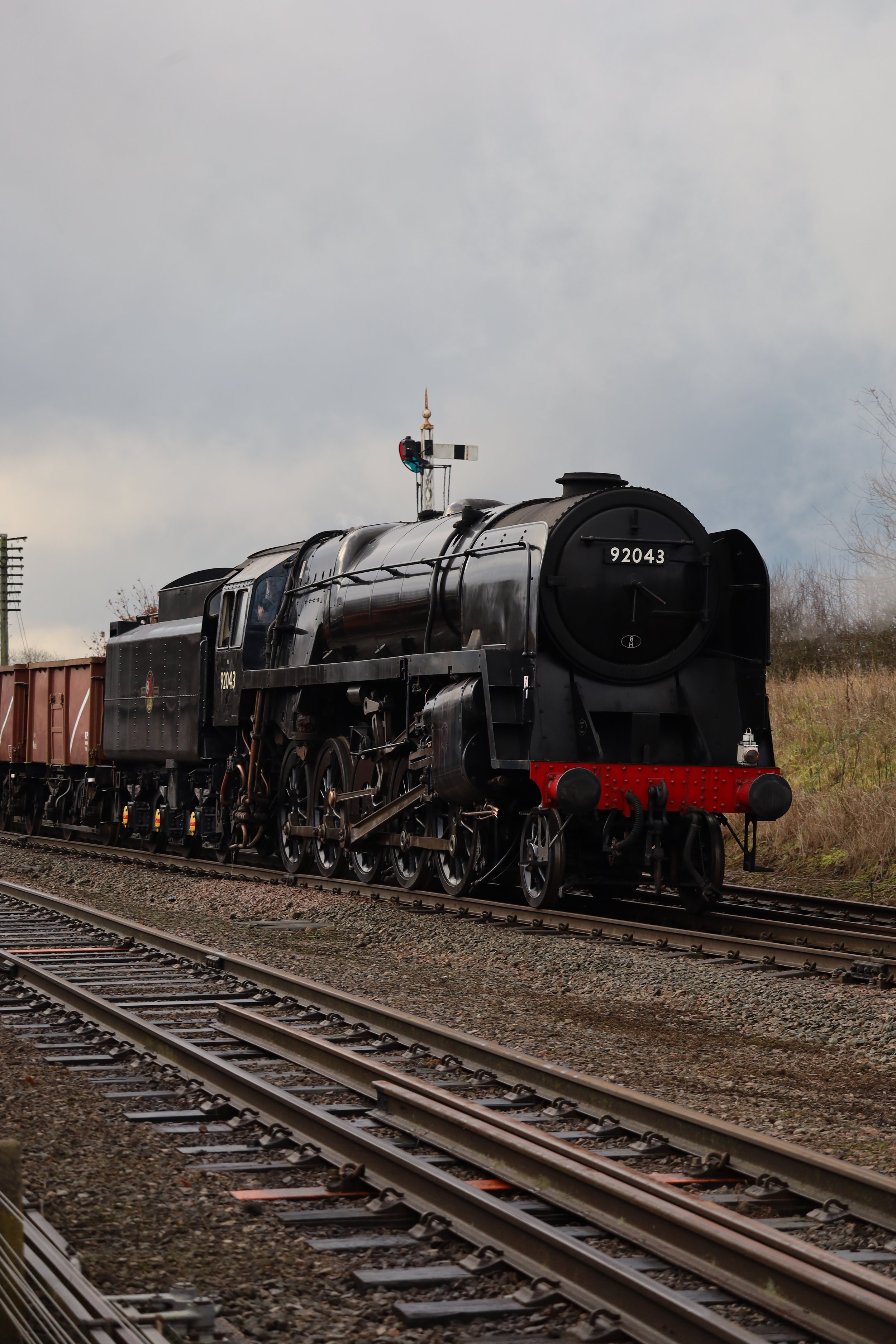 A black steam locomotive train with the number 92043 on the front, moving on railway tracks under a cloudy sky.