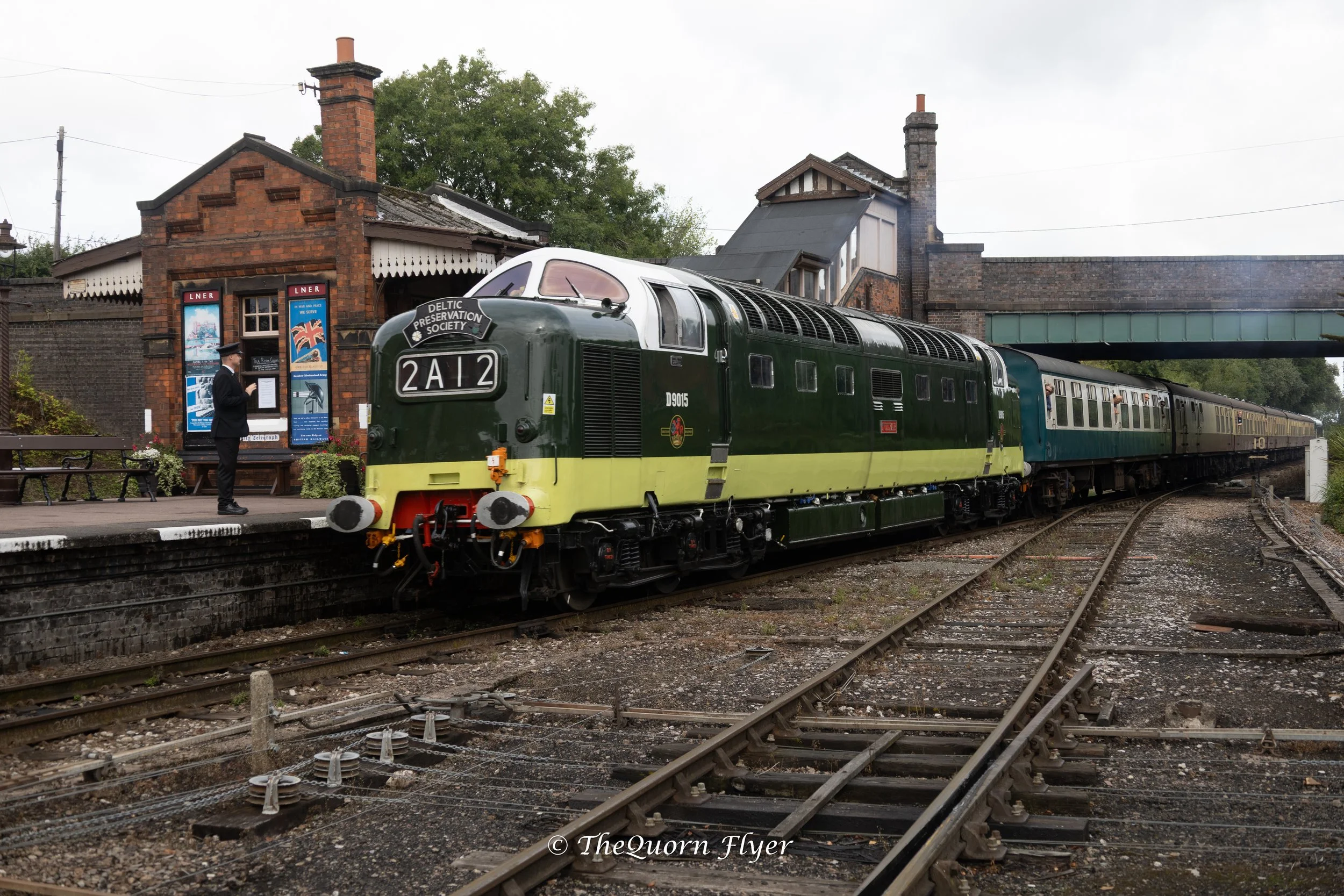 A vintage green and cream train at a train station platform with an older brick building and a man in a train conductor's uniform reading a schedule or announcement. The background includes trees and an overcast sky.