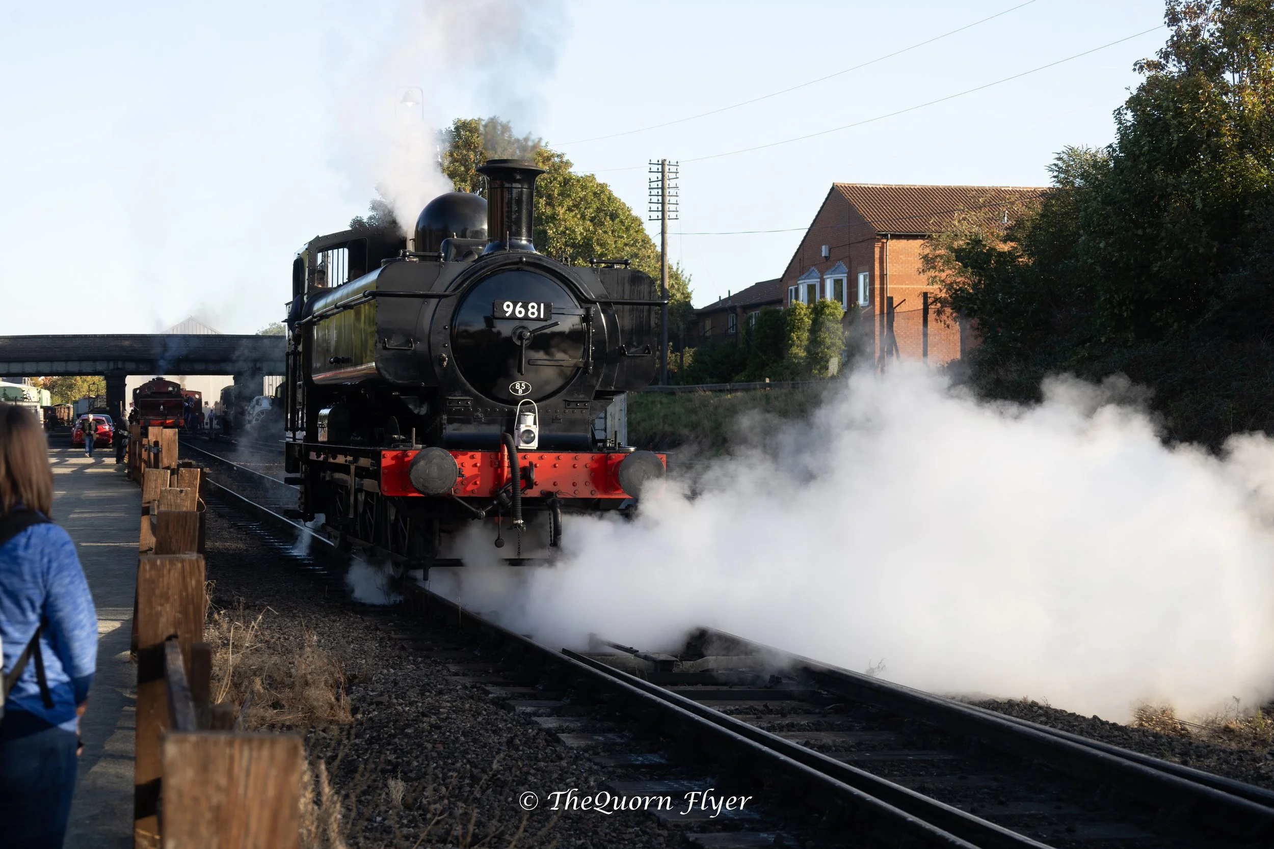 A vintage black steam locomotive train pulling into a station with smoke billowing from its chimney, while people stand nearby and a building with a brick exterior is visible in the background.