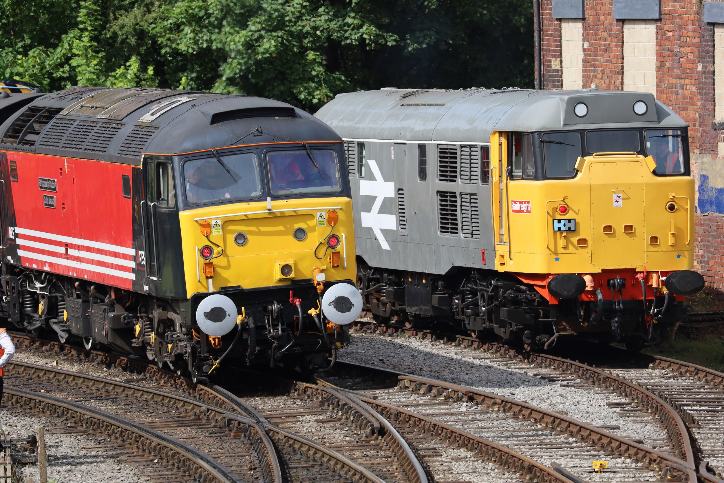 Two train engines on railway tracks, with a brick building and green trees in the background.