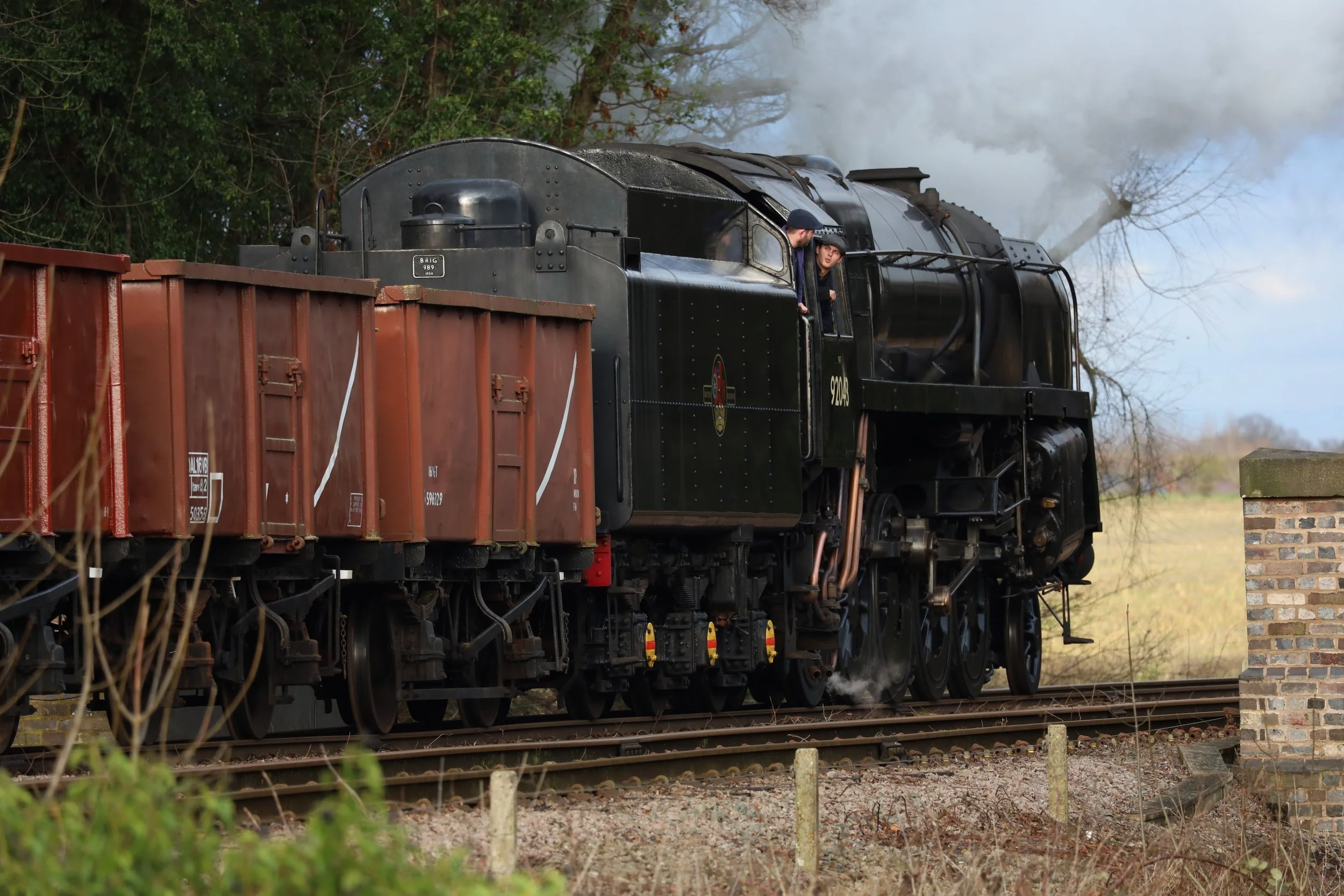 A vintage black steam train moving on railway tracks with two men visible in the locomotive's cab, smoke billowing from the chimney, surrounded by trees and under a cloudy sky.