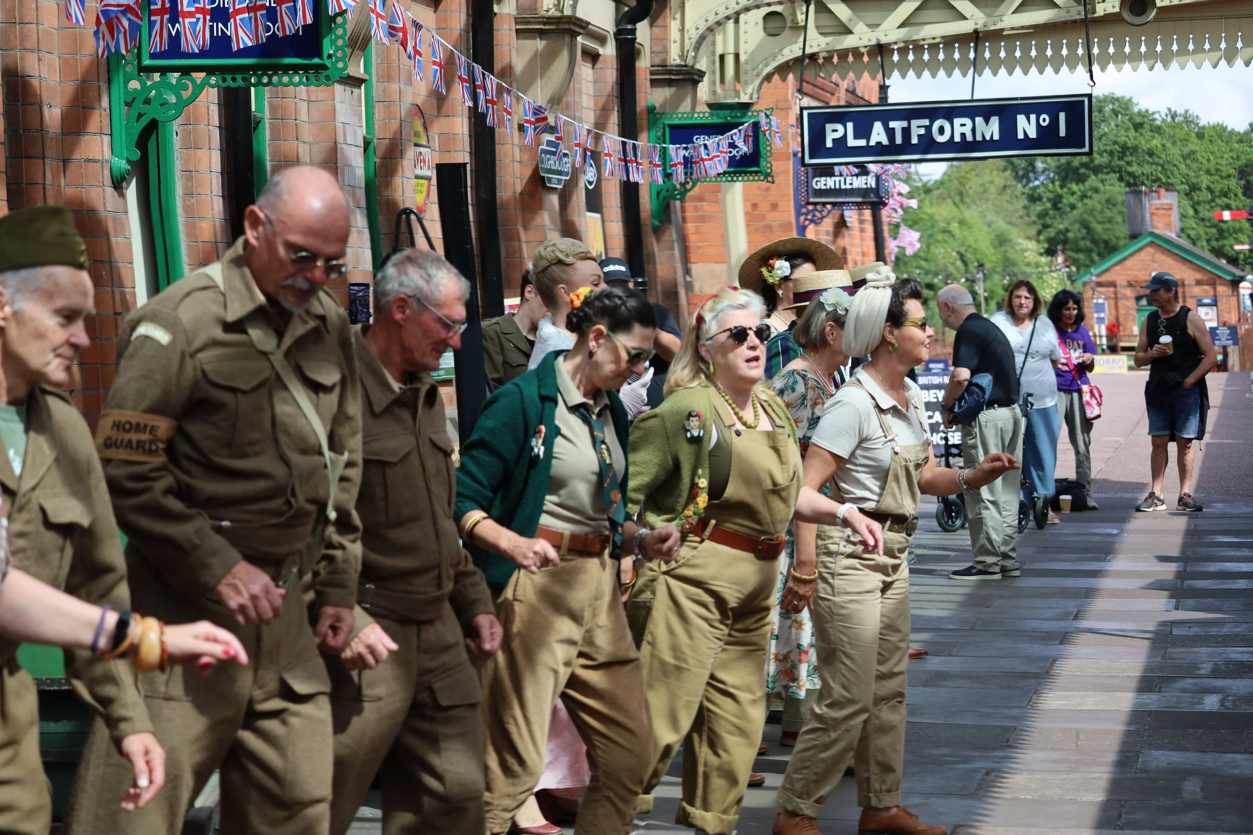 A group of people dressed as military veterans standing in a line outdoors near a brick building with Union Jack flags and signs that say 'Platform No 1', and other vintage-style decorations.