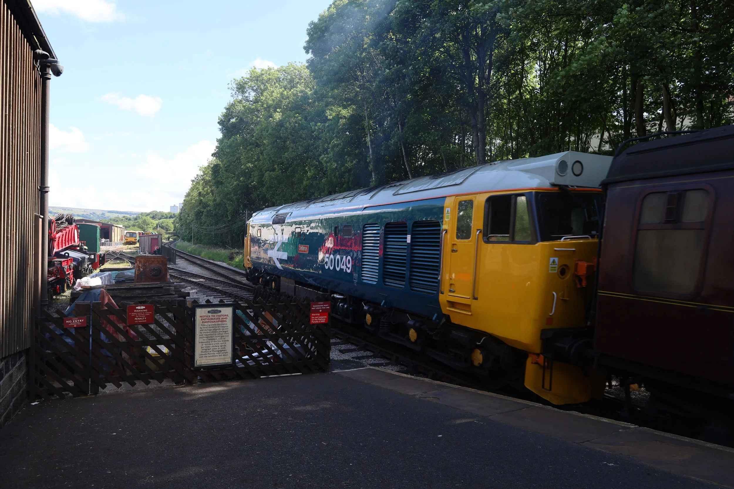 A passenger train passing through a level crossing with barriers and warning signs, surrounded by green trees on a sunny day.