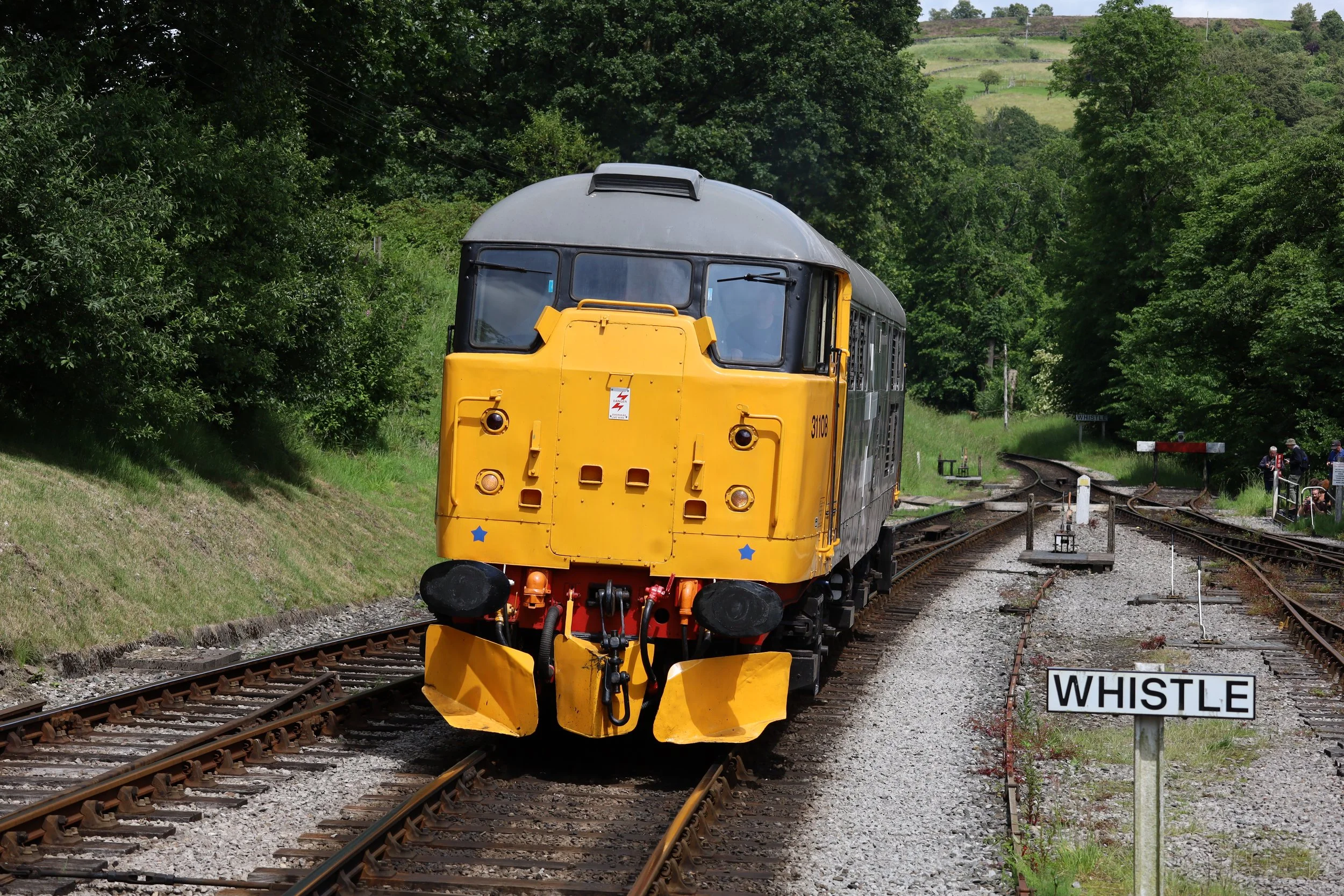 A yellow and black train on railway tracks surrounded by green trees and hills, with a sign that reads 'WHISTLE' nearby.