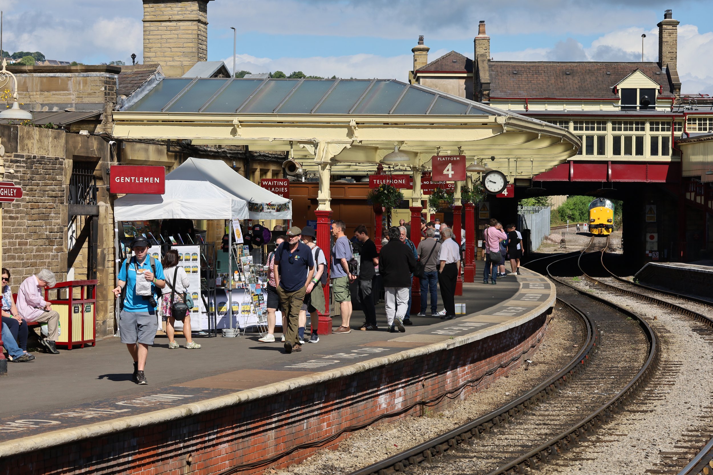 A bustling train station platform with people waiting, walking, and shopping. The platform features a red sign labeled 'Platform 4,' a round clock, and a small tent with souvenirs. A lowered train track is visible with an approaching yellow train in the background. The station has vintage architecture with a canopy and brick walls, under a partly cloudy sky.