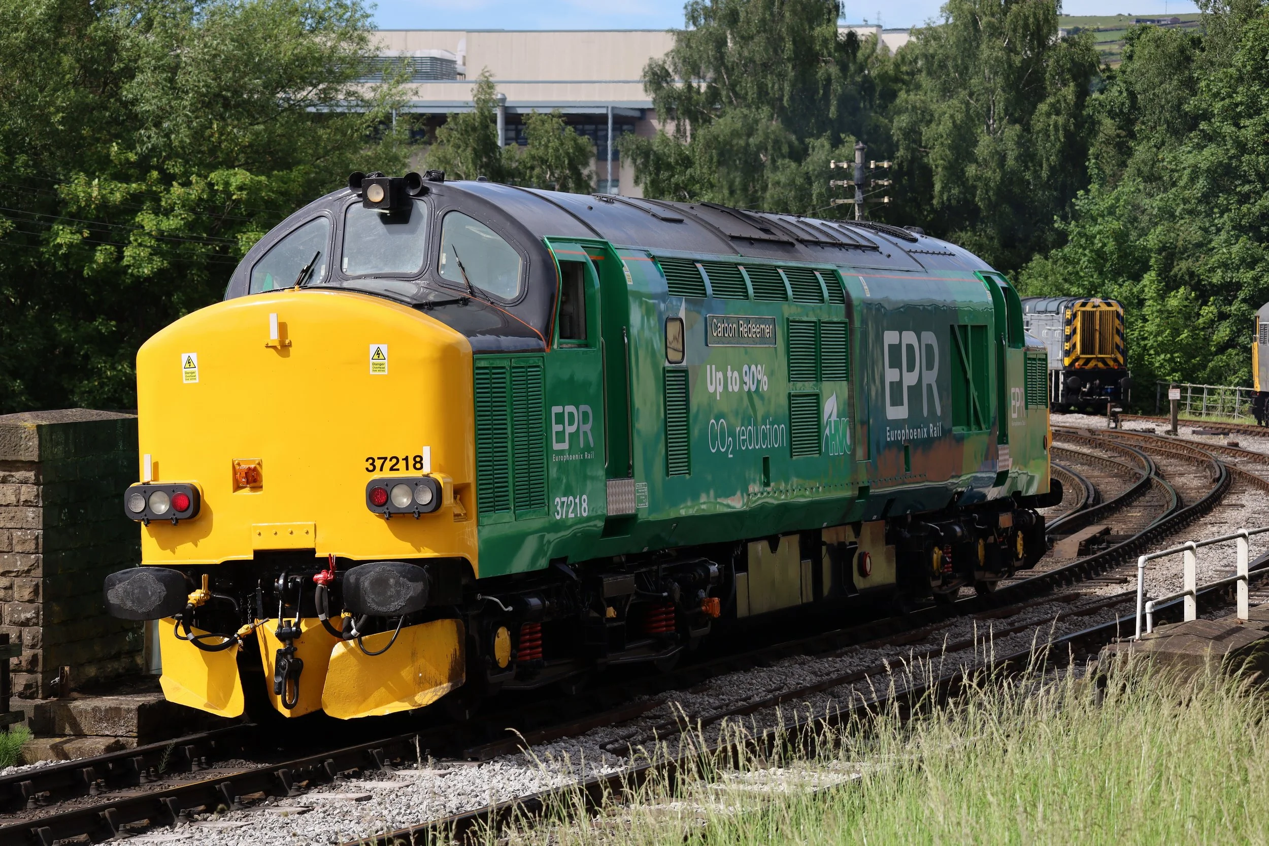 A green and yellow train labeled 'Carbon Reducer' on a railway track with trees and a building in the background.