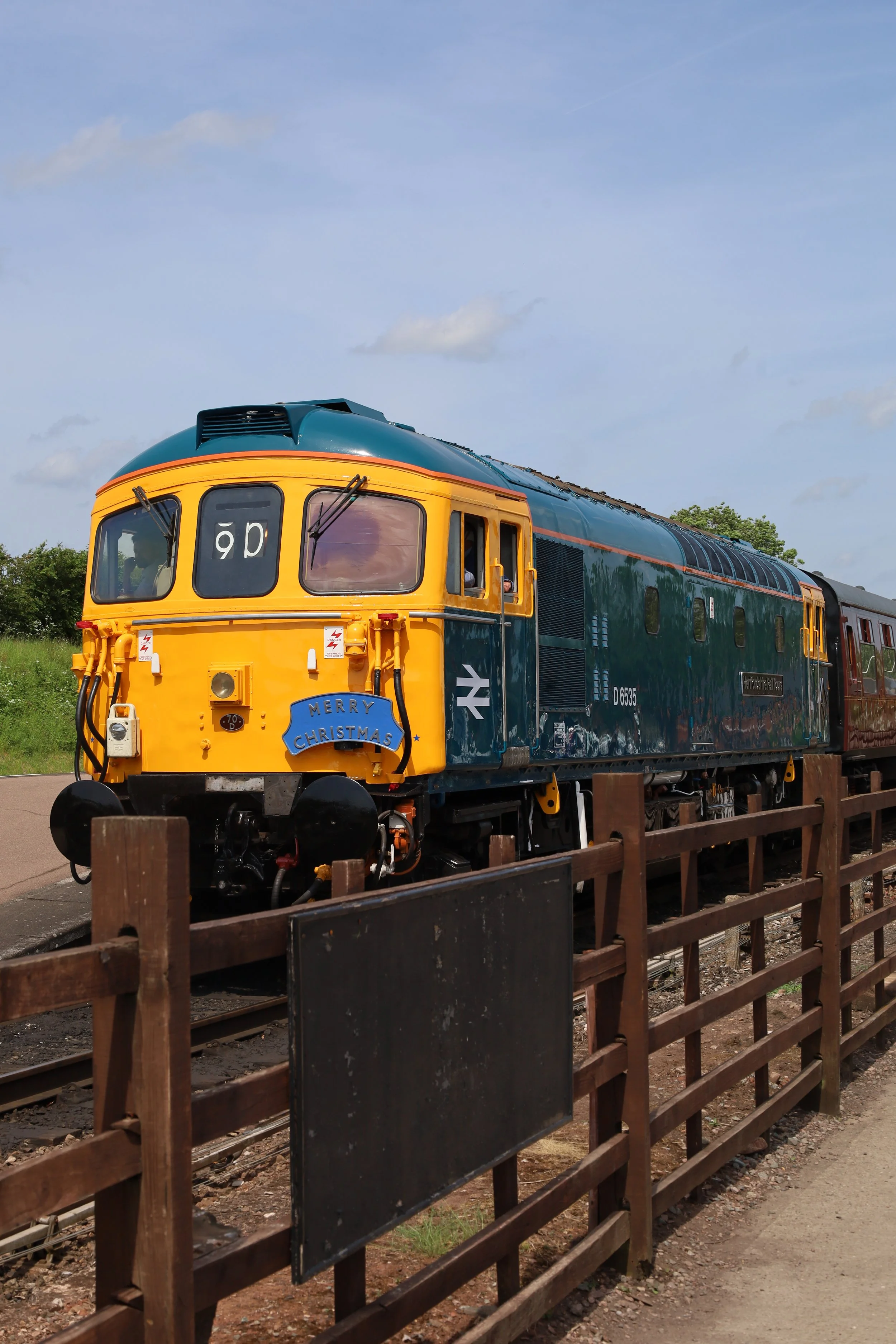 Colorful train with yellow front, blue roof, and dark blue body traveling on tracks, with a wooden fence in the foreground, green trees, and a partly cloudy sky in the background.