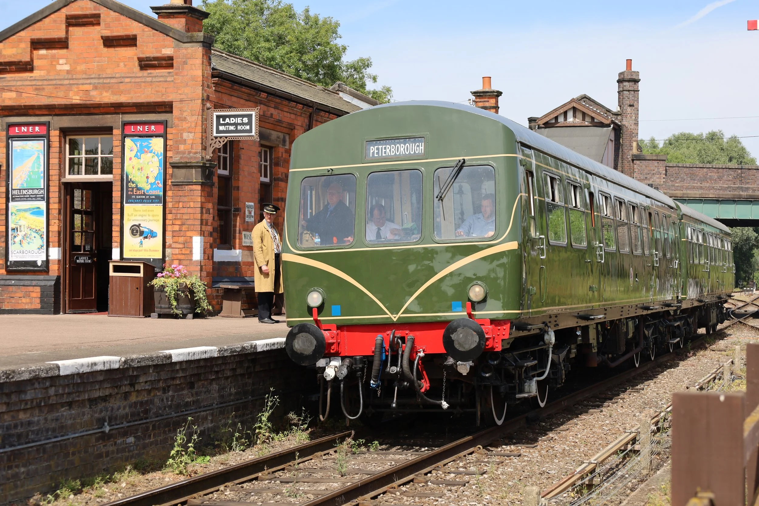 A vintage green train on a railway platform with a sign reading 'Peterborough.' A station staff member is standing next to the train, and there is a station building with posters and signs, including one for a ladies' waiting room.