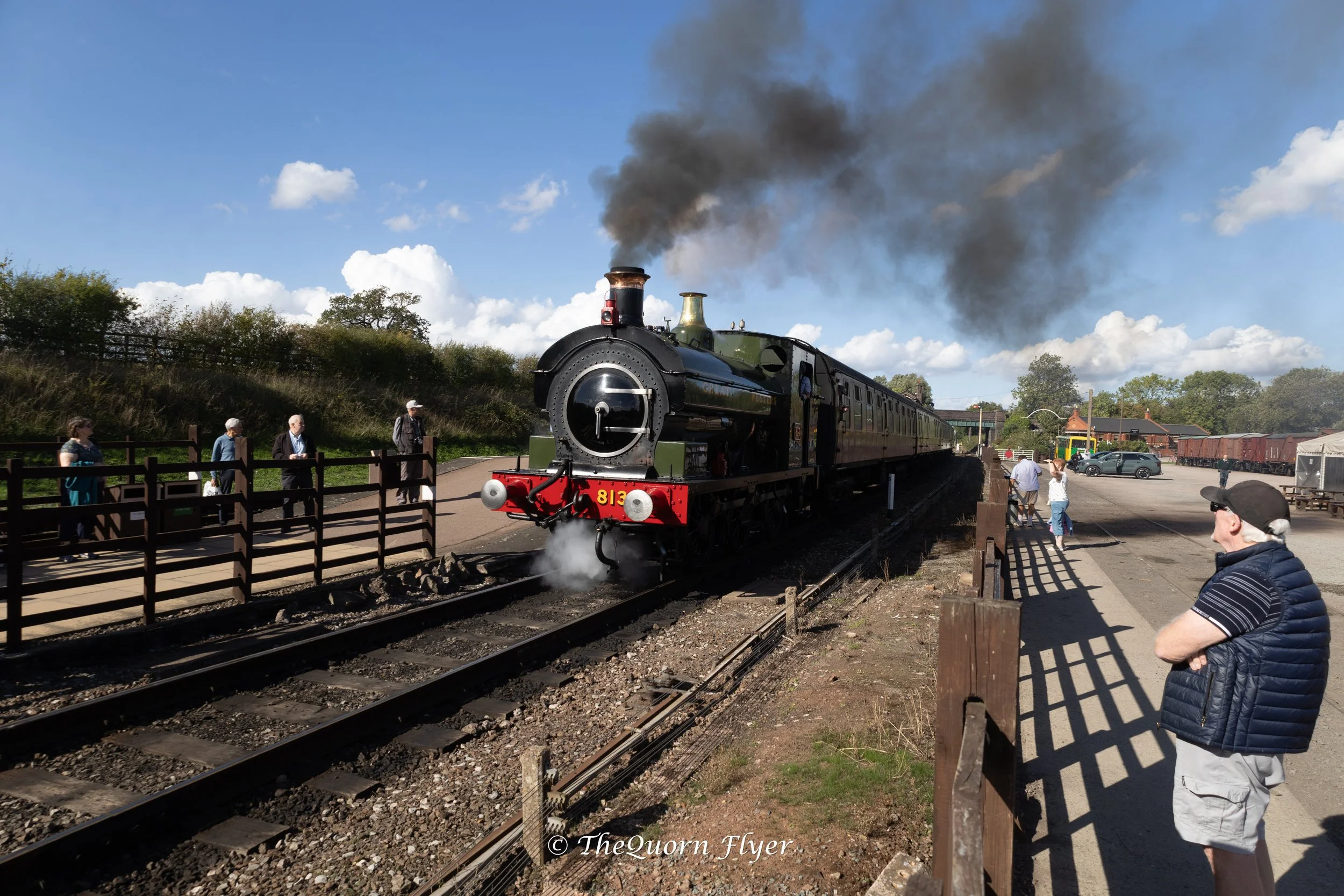 A vintage steam train with black and green colors traveling on railway tracks, emitting black smoke into a blue sky with white clouds, while people stand behind a wooden fence watching the train pass by.
